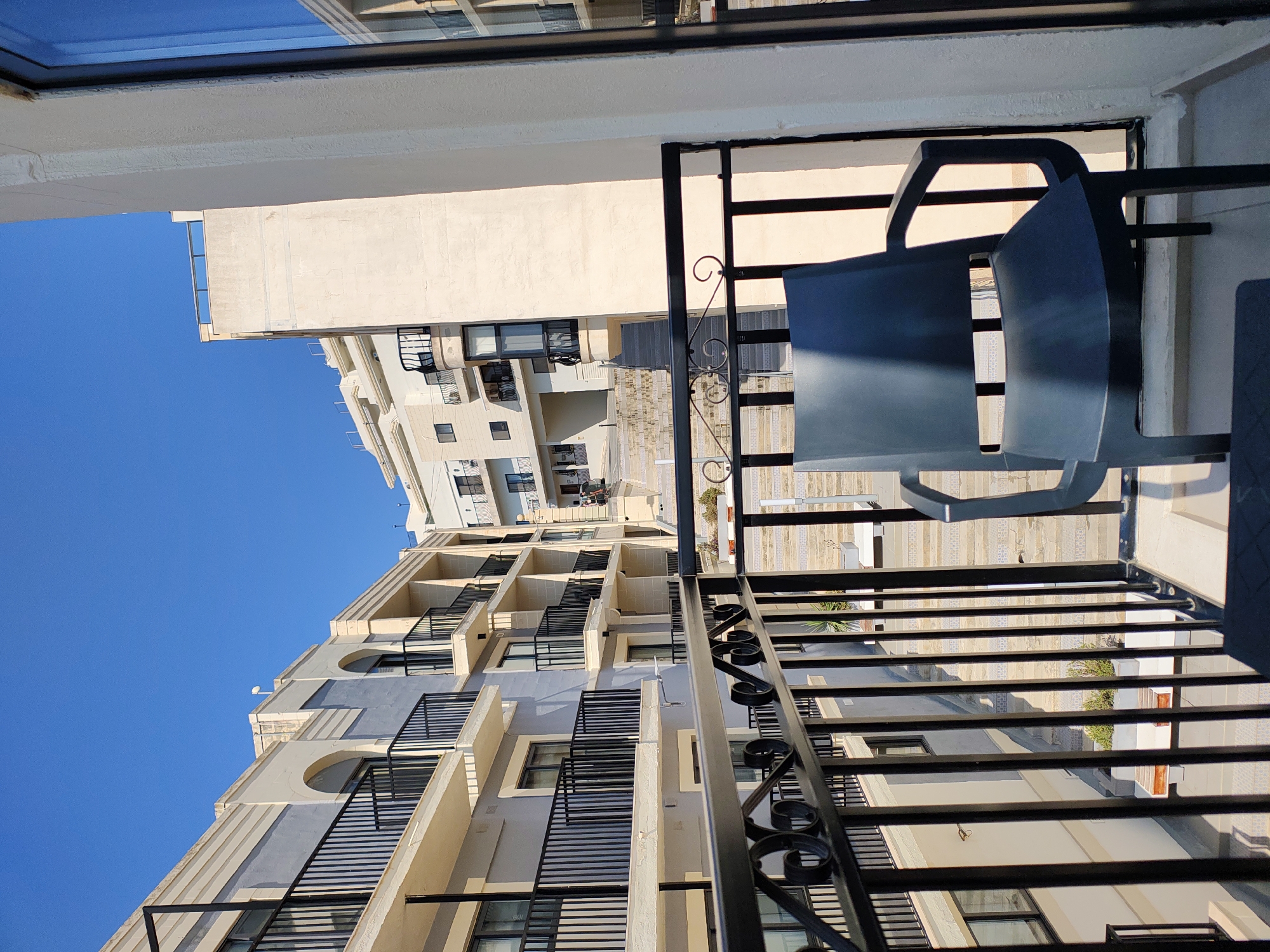 The image depicts a view from a balcony overlooking an urban landscape in Manikata, Malta. The foreground features a dark blue plastic chair placed on the balcony, with a decorative black metal railing in front of it. Beyond the railing, a complex of multi-story buildings with balconies and windows is visible. The architecture appears to be modern Mediterranean, with light-colored facades and dark railings. A set of outdoor stairs is also visible, leading down between the buildings. The sky is a clear, bright blue. The scene suggests a residential or hotel complex in a sunny climate.