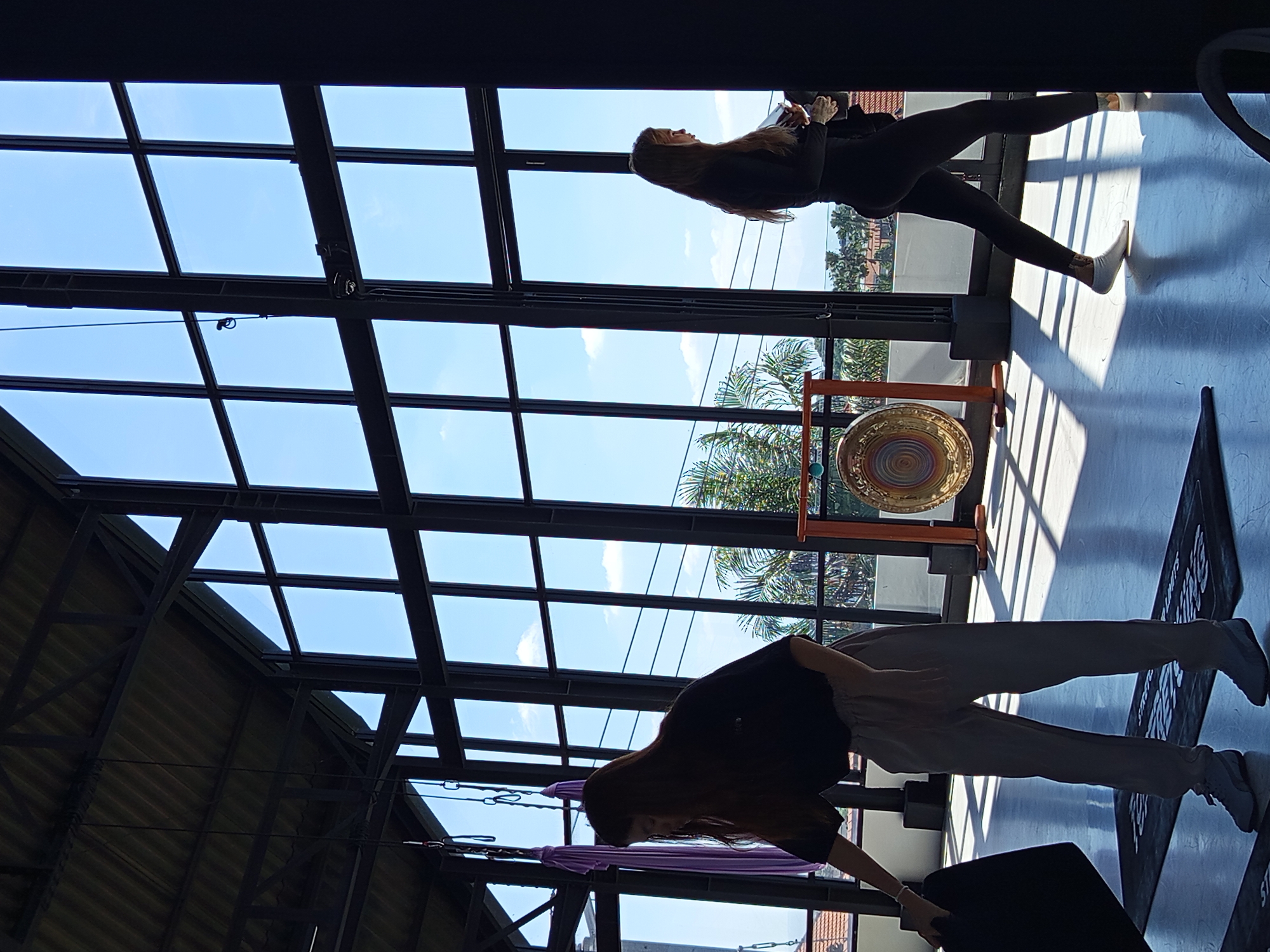 The photo depicts an interior space with a grid-patterned glass ceiling showing a bright blue sky with scattered clouds, taken in North Kuta, Indonesia. Two figures are seen: one positioned horizontally in the upper right and another oriented diagonally in the lower center. The person in the upper right is wearing dark, form-fitting clothing and light-colored footwear. The person in the lower center is dressed in a dark top, loose, light-colored pants, and sneakers. There is a golden gong on a wooden stand visible in the mid-ground. A purple aerial hammock is suspended from the ceiling. A mat with text is visible on the floor.