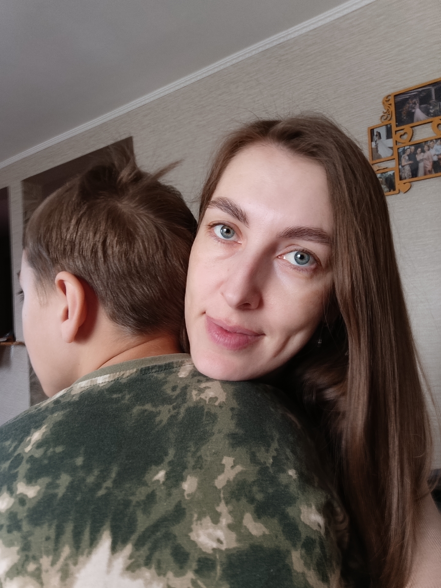 A woman with long brown hair and blue eyes smiles at the camera, her head resting on the shoulder of a boy with short brown hair wearing a green and white camouflage shirt. They are indoors, with a wall featuring a photo collage in the background. There is a television on the wall as well. The location is likely indoors in Фоки, Russia, based on prior knowledge.