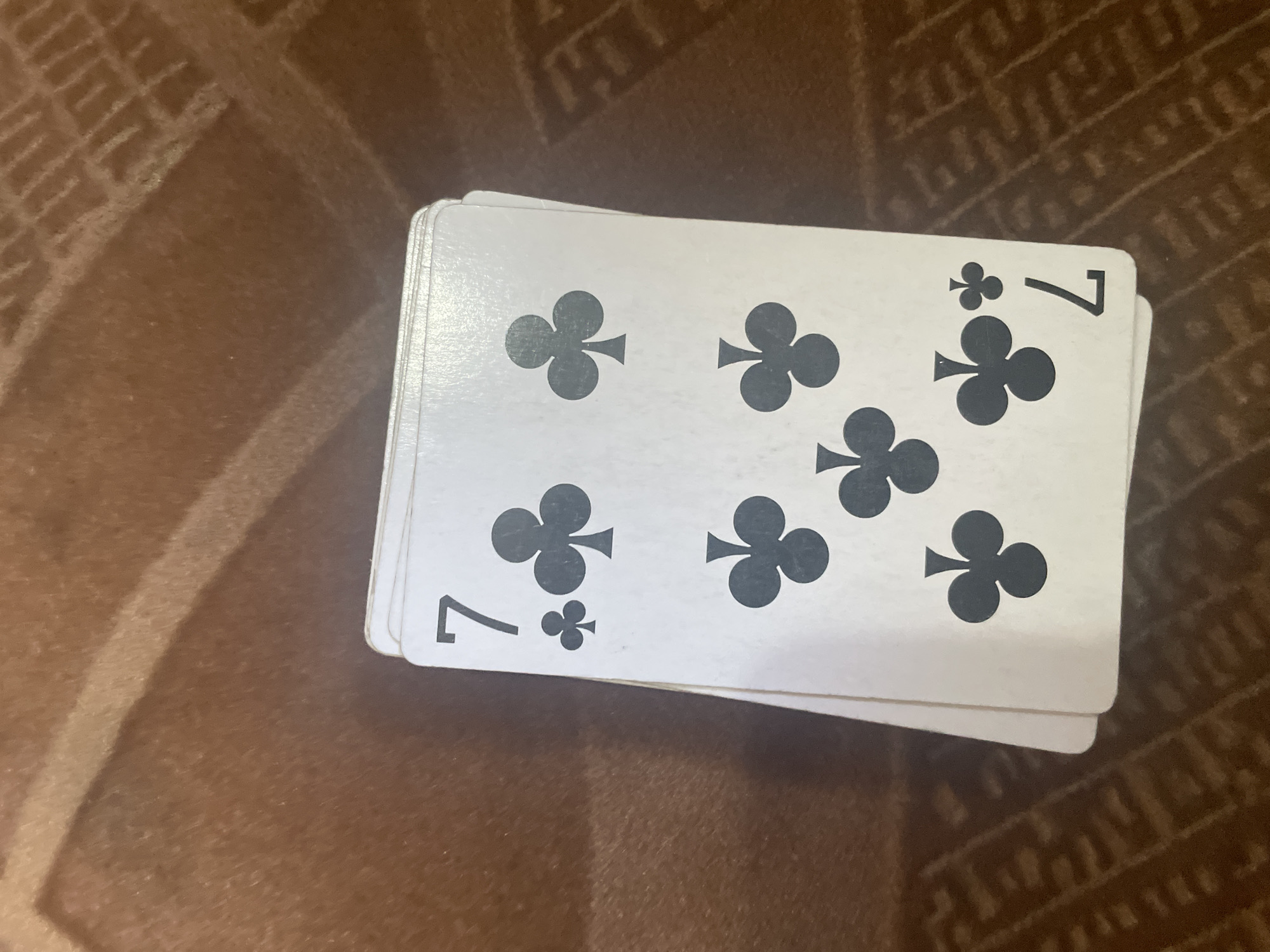 The image shows a deck of playing cards, specifically the Seven of Clubs, fanned out on a patterned brown surface. The cards are white with black pips and numbers. The top card clearly displays a "7" in the top left and bottom right corners, with multiple club symbols scattered across its face. The other cards in the deck are partially visible, suggesting they are also Seven of Clubs cards, as indicated by the visible club symbols. The background surface has a textured pattern. The lighting suggests it is indoors, with no indication of time of day or weather. The image does not depict people or any specific activity or event beyond the arrangement of the cards. The location context provided is Esik, Kazakhstan.