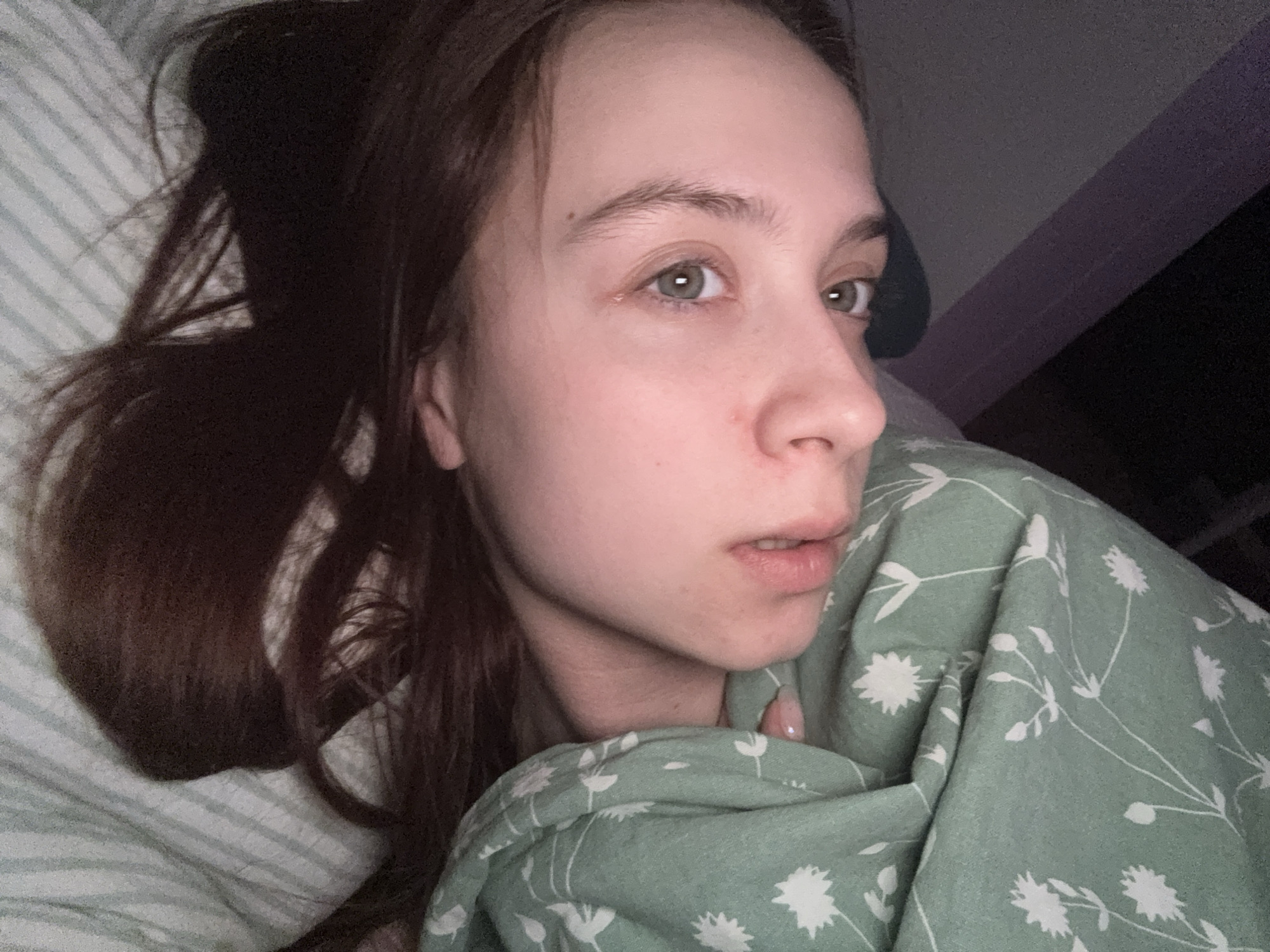 A young woman with light skin and brown hair is lying in bed. She is partially covered by a green blanket with a white floral pattern. The bedding beneath her is striped. The environment appears to be a dimly lit bedroom, suggesting it could be night or a room with limited natural light. It is not possible to determine the location in the city of Прикамский, Russia based on the image alone.