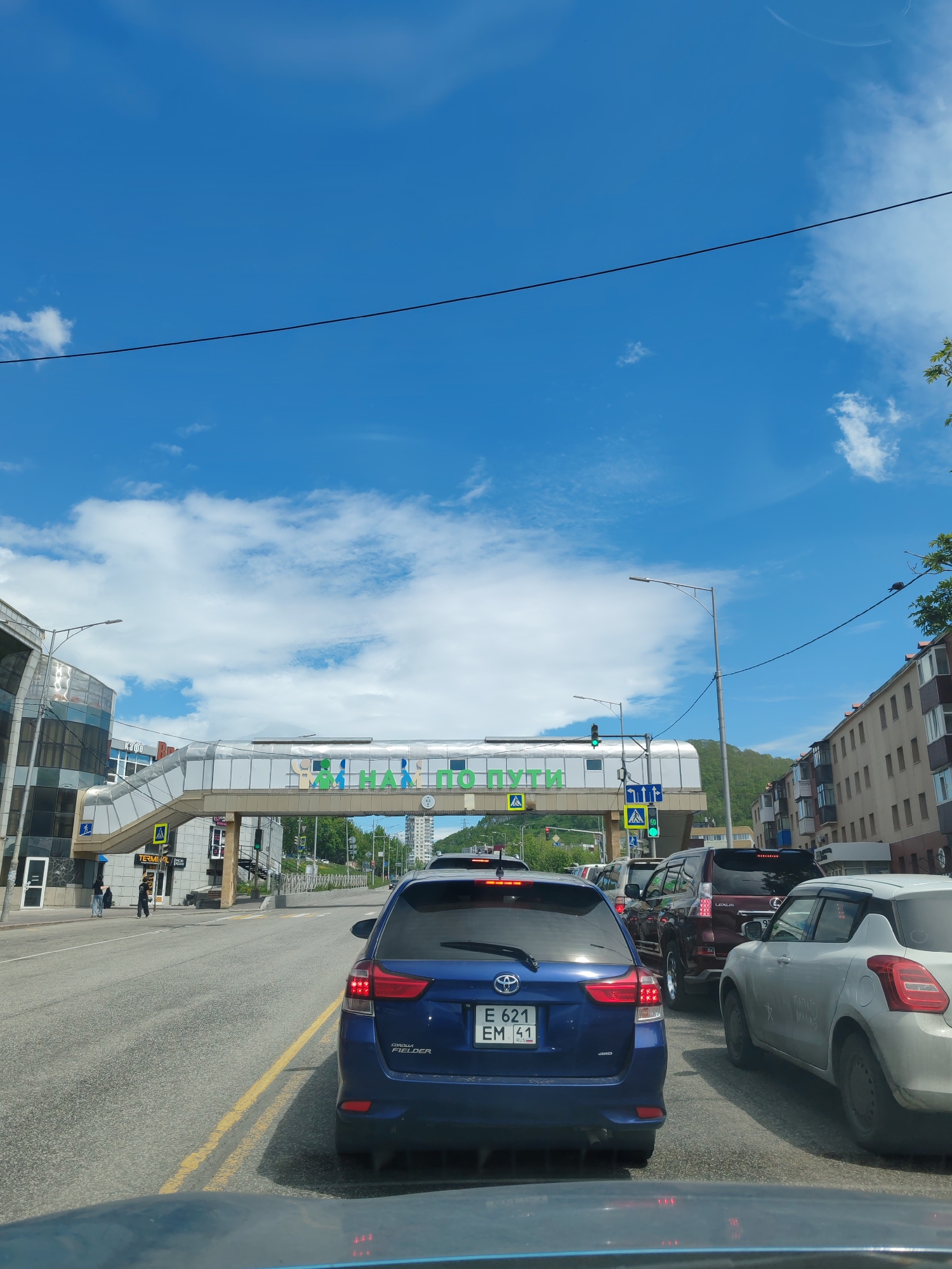 The image captures a street scene in Petropavlovsk-Kamchatsky, Russia, on a clear day with blue skies and scattered clouds. In the foreground, the rear of a blue Toyota Corolla Fielder is visible, facing away from the camera, with its license plate reading "E 621 EM 41 RUS". To the right, a white Suzuki Swift is parked.
The street is lined with cars, suggesting a traffic jam or a busy intersection. A pedestrian overpass spans the road, adorned with green text that reads "СИГНАЛ ПО ПУТИ" (Signal en route). This overpass connects two buildings: on the left, a modern building with a glass facade, and on the right, a multi-story residential building. Traffic lights and street lamps are also present. In the background, lush green hills rise, characteristic of the Kamchatka region.
A wire stretches across the sky, with a pigeon perched on it in the upper right corner of the image. The overall impression is of a typical urban environment in a city known for its dramatic natural landscape.