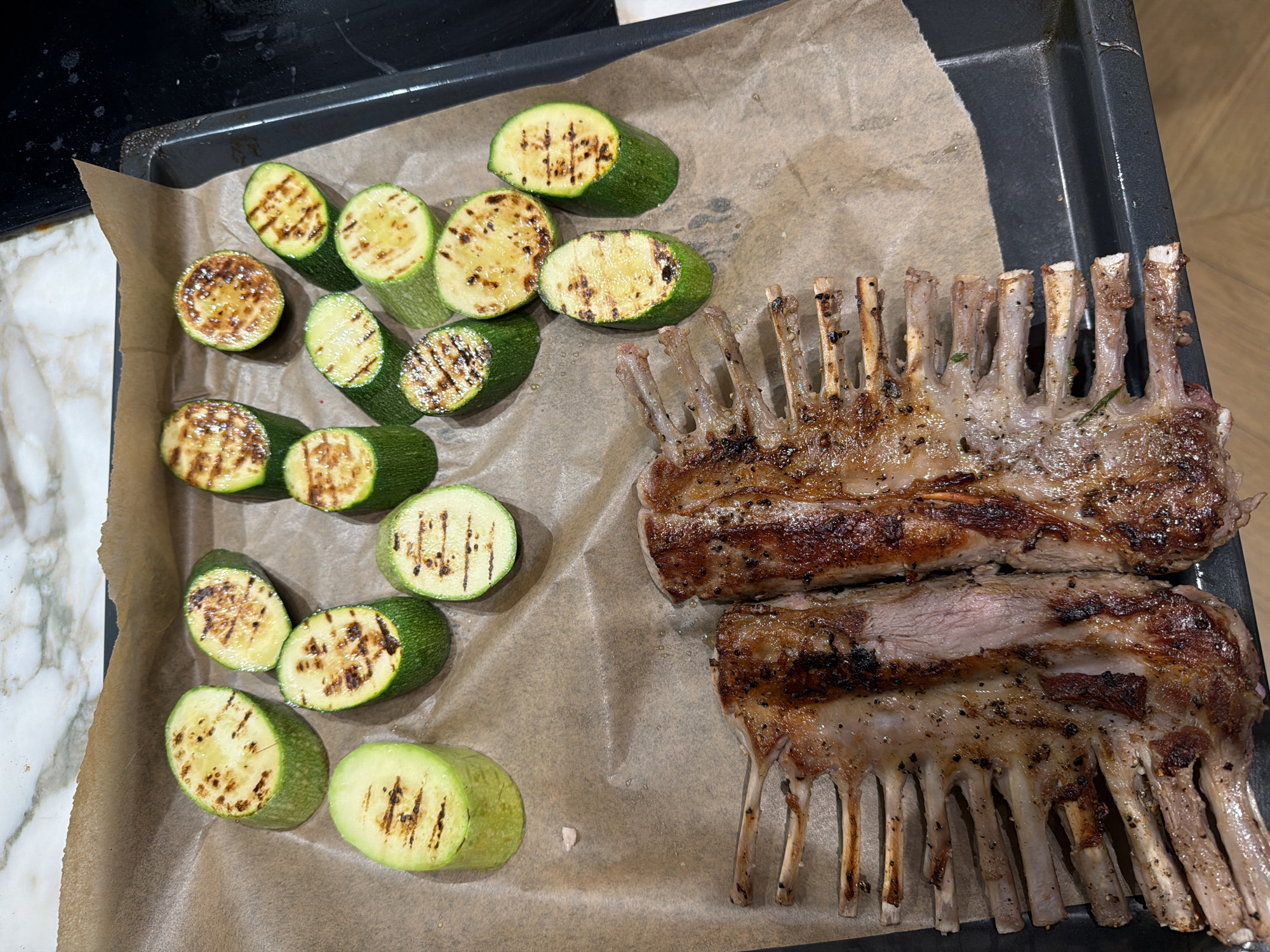 Two racks of lamb, seasoned with black pepper and herbs, are presented on a baking sheet lined with parchment paper, alongside several grilled zucchini rounds. The lamb exhibits a rich, seared exterior with visible grill marks and a pink, medium-rare interior. The zucchini slices are also marked by the grill, with some showing a slightly softened texture. The baking sheet is positioned on a tiled surface, potentially in a kitchen setting. There is no indication of a specific location in Moscow, Russia.