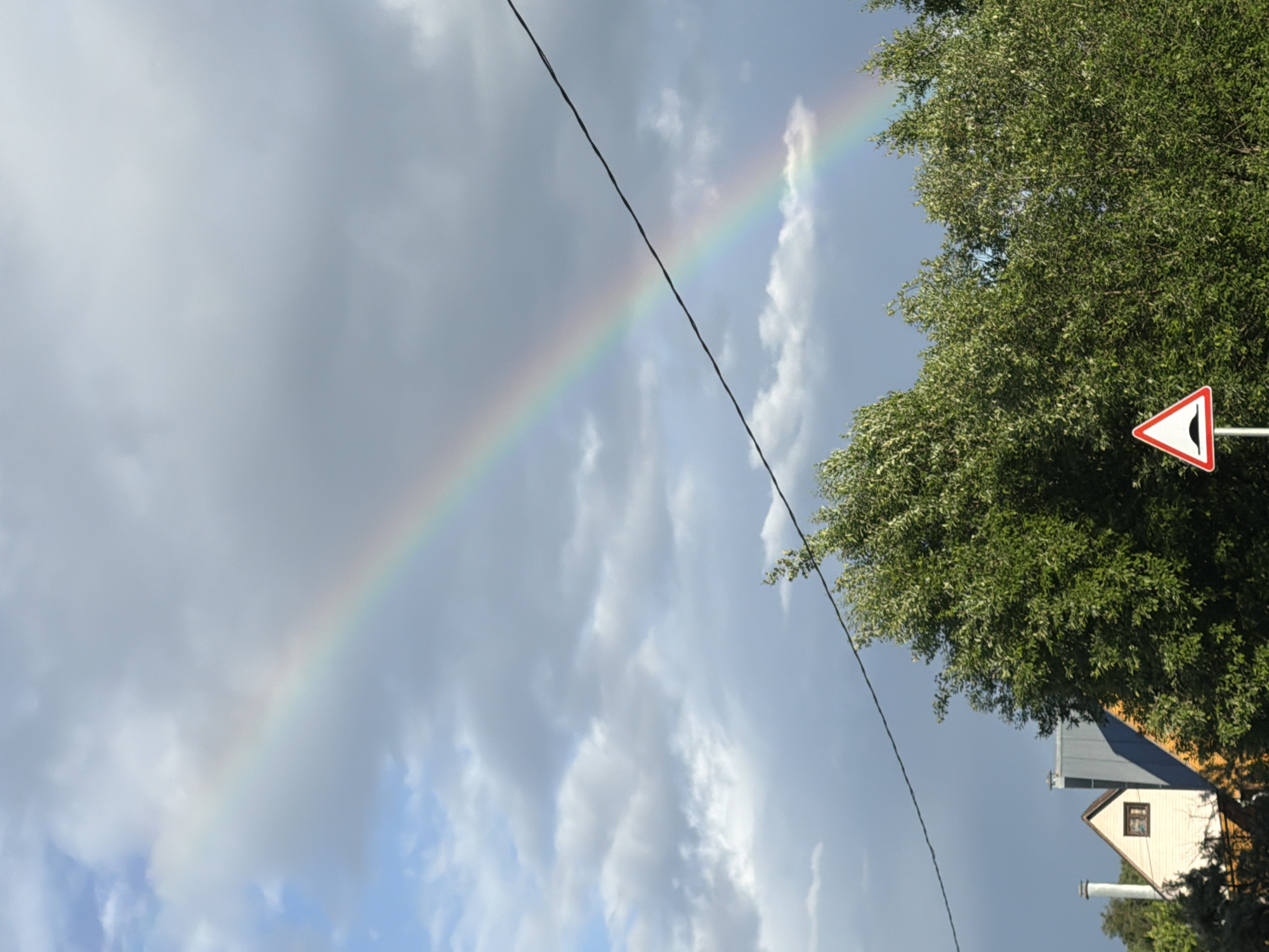 The image depicts a rural scene in Satino-Russkoe, Russia, featuring a vibrant rainbow arching across a cloudy sky. In the foreground, lush green trees dominate the lower portion of the frame. A portion of a house with a pitched roof and a small window is visible on the left, peeking through the foliage. To the right of the house, a prominent traffic sign indicating a speed bump is mounted on a pole. A thin black wire stretches diagonally across the upper portion of the image, intersecting the rainbow and the trees. The sky is a mix of blue and grey clouds, with patches of sunlight breaking through, illuminating the rainbow. The overall impression is one of a post-rainy, atmospheric landscape.