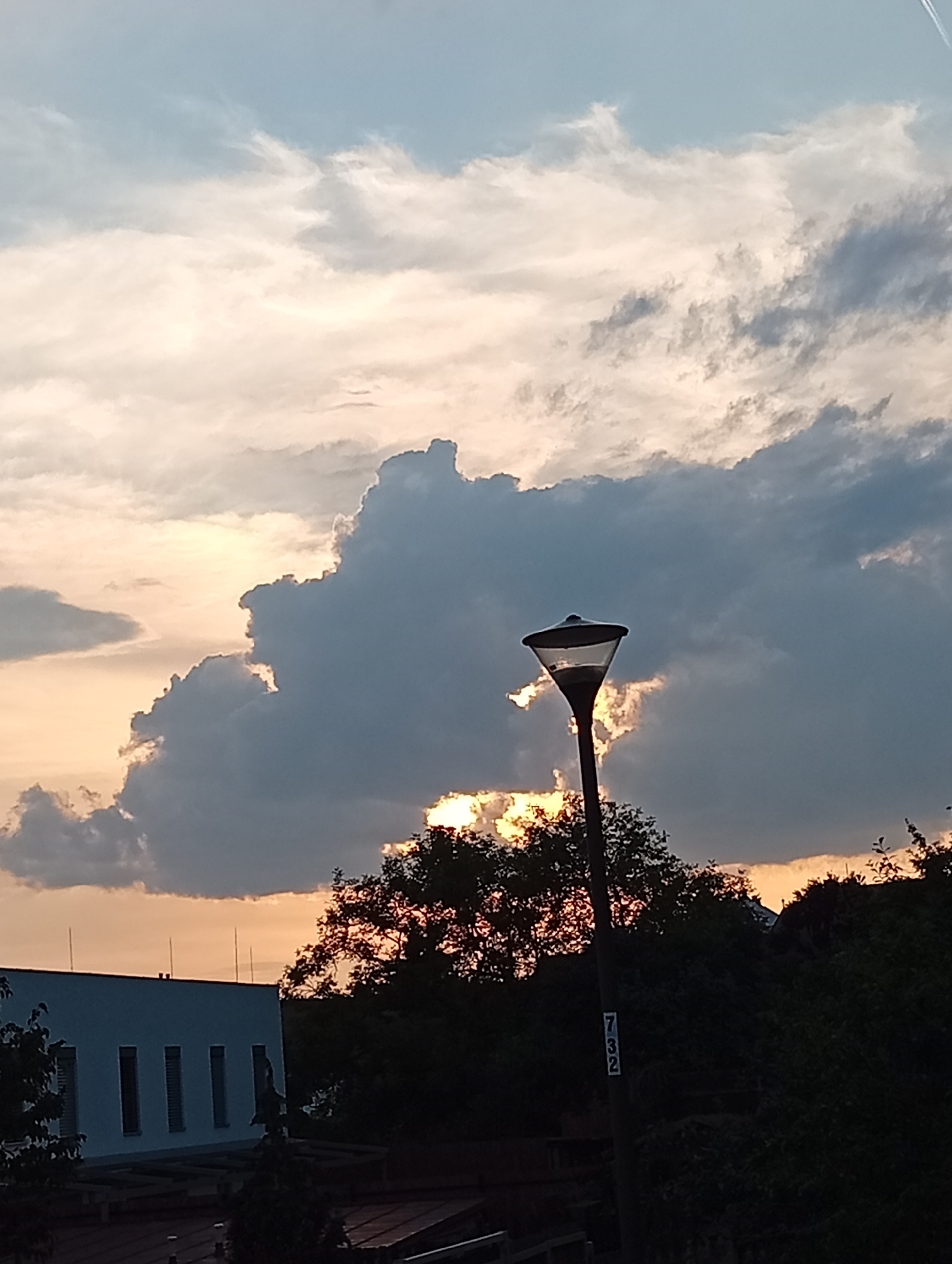 The image depicts a cloudy sky over the city of Tišnov, Czechia, at dusk. The sky is a mixture of light blue, white, and dark gray clouds, with the setting sun casting a warm, orange glow through a gap in the clouds. In the foreground, a modern-style street lamp with a transparent shade stands tall. Behind the lamp, silhouetted trees and the roofline of a building are visible. To the left, a white building with a flat roof and evenly spaced windows or ventilation openings can be seen. The overall scene is one of a serene evening in an urban environment.