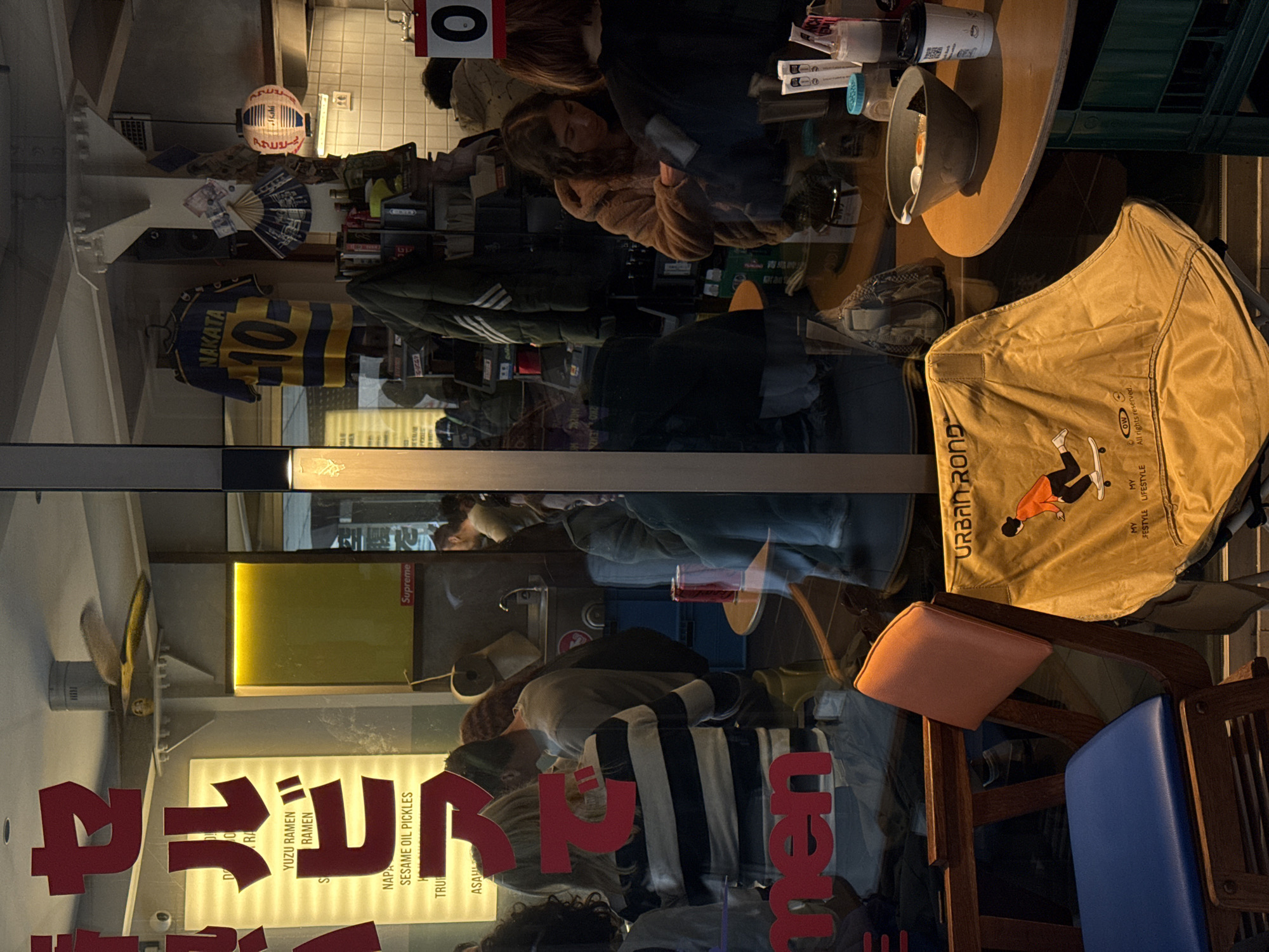 The image appears to be a reflection in a storefront window. The window displays the word "RAMEN" in large red characters, along with text in Japanese and English. Behind the window are a variety of objects, including a soccer jersey with "NAKATA 10" printed on it, a round table, and a tan bag labeled "URBAN ROND." Seated at the table are several people. There are also chairs with wooden frames and blue and pink cushions. The overall scene suggests an urban restaurant setting. The text "青島啤" suggests the presence of a Chinese restaurant and beer. "セルビア" is in the upper-left, confirming the location in Serbia, which means the "青島啤" text is a reference to Tsingtao beer, a popular brand. "ASAHI" can also be seen, referring to Asahi beer.