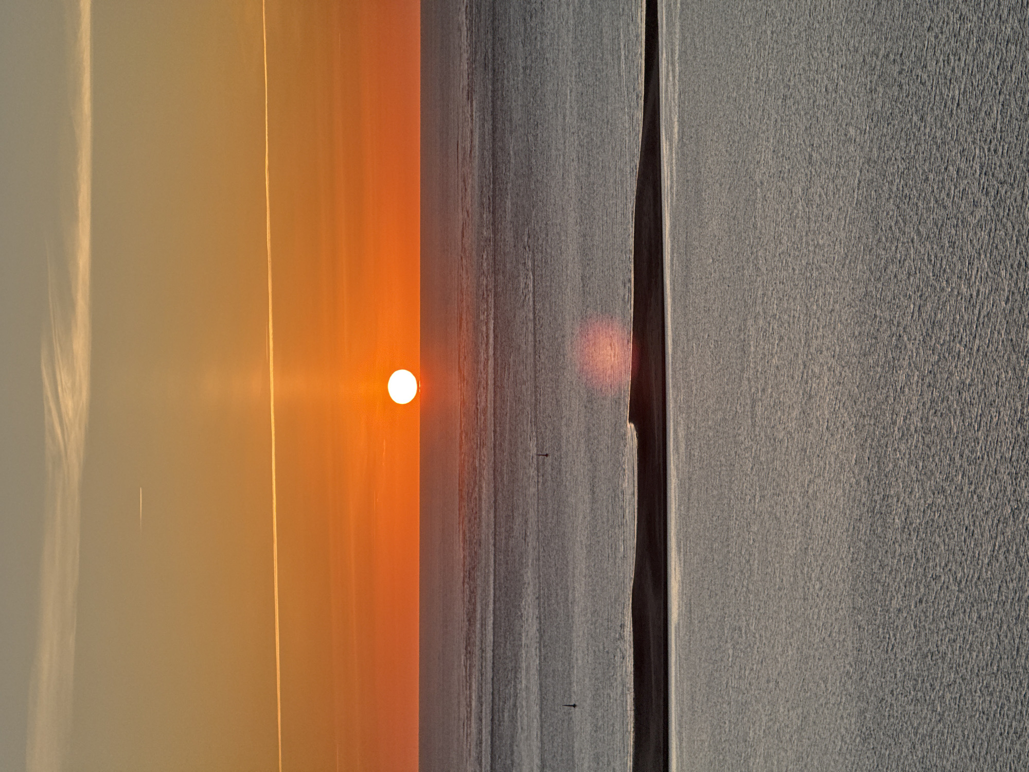 This is a vertical, eye-level, full shot of a sunset over the ocean, likely the Atlantic Ocean off the coast of La Teste-de-Buch, France. The sun, a bright orange disc, is positioned just above the horizon, casting a vibrant orange glow across the sky and a shimmering reflection on the water. The sky transitions from a pale yellow near the horizon to a dusty blue at the top, with thin, wispy clouds and two distinct contrails from aircraft visible.
The ocean is divided into three distinct horizontal bands. The uppermost band, closest to the horizon, is a calm, dark gray expanse with subtle ripples. Two small, dark buoys are visible in this section. Below this is a slightly lighter gray band, also calm, with a faint reflection of the sun's light. The lowest band, occupying the bottom half of the image, is a muted gray, textured with small, choppy waves that catch the light.
In the foreground, a dark, low-lying landmass, possibly sand dunes or a very low island, separates the upper two water bands from the bottom one. Its surface is mostly in shadow, but a thin line of bright light runs along its top edge, reflecting the setting sun. The overall atmosphere is serene and tranquil, typical of a sunset scene over a coastal environment. There are no people or any other discernible subjects or actions within the frame.
