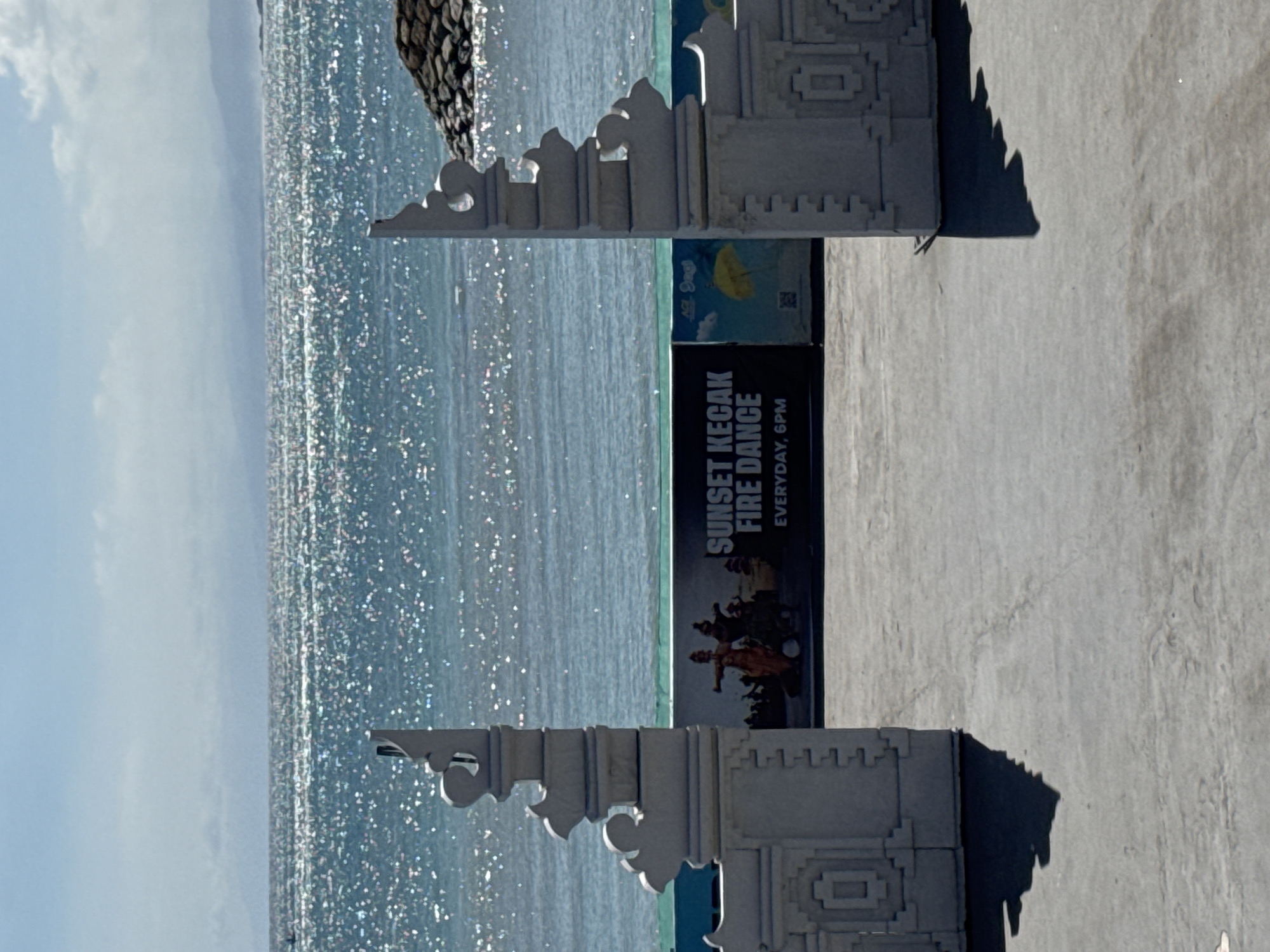 The image depicts a view of the ocean in Kuta, Indonesia. A sign advertising "Sunset Kecak Fire Dance, Everyday, 6PM" is positioned in the foreground, framed by ornate, white stone carvings on either side. The ocean extends towards the horizon, shimmering with reflected sunlight. The sky is partially cloudy.