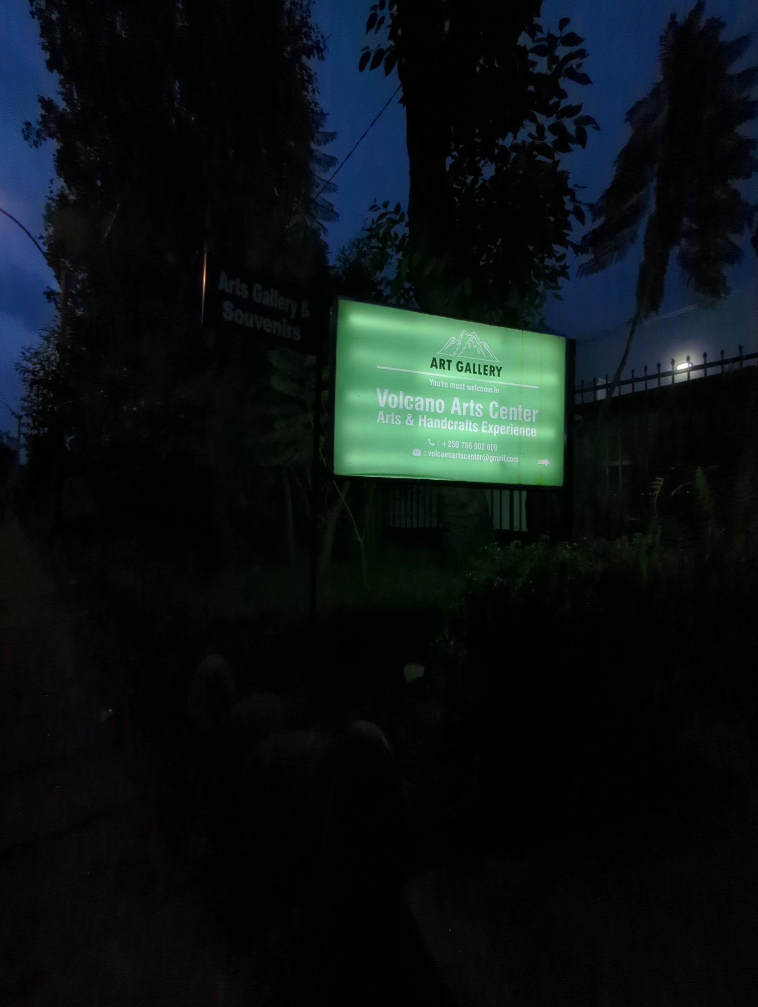 The image depicts a dimly lit scene at night, likely in Musanze District, Rwanda. A bright, illuminated green sign is the central focus, advertising "ART GALLERY" with the tagline "You're most welcome in Volcano Arts Center Arts & Handicrafts Experience." Below this, contact information is visible: a phone number "+250 786 902 609" and an email address "volcanoartscenter@gmail.com". The sign also features a graphic of mountains. To the left of this main sign, a smaller, darker sign reads "Arts Gallery & Souvenirs". Surrounding the signs are dark silhouettes of trees, and a fence with lights can be seen in the background. The overall impression is of an establishment operating at night, with its signage prominently lit.