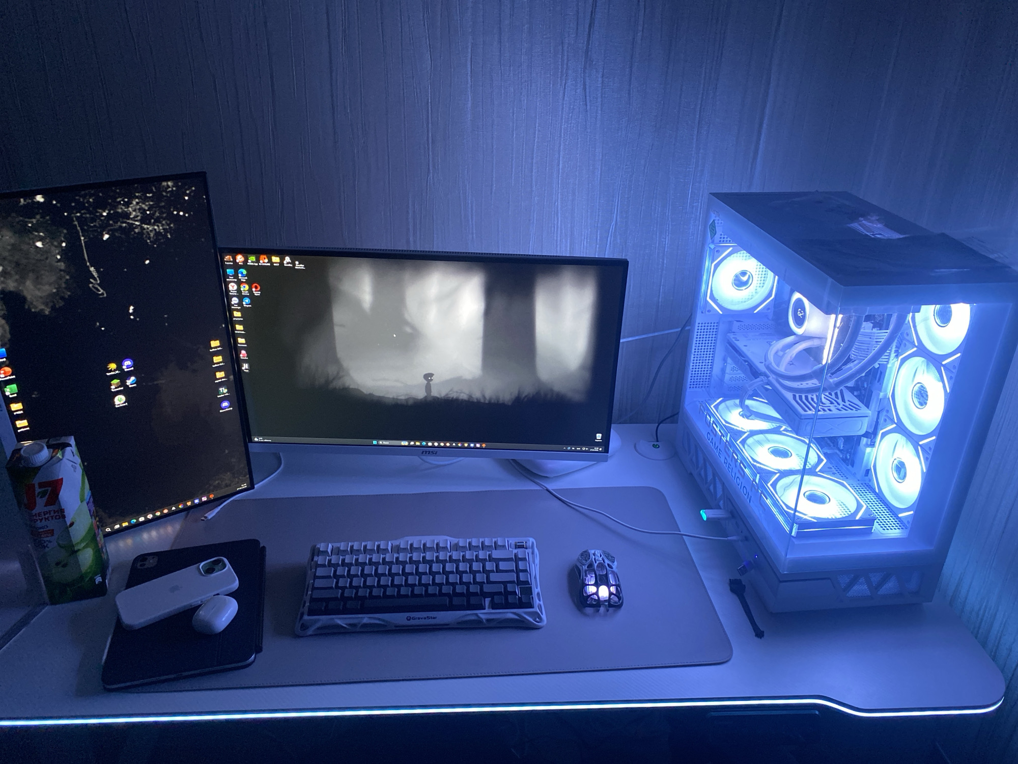 This image displays a modern computer workstation, likely a gaming setup, illuminated by cool blue and cyan LED lighting, suggesting an evening or nighttime setting. The room features textured, vertically striped wallpaper in a dark blue or grey-blue hue.
The setup includes two computer monitors. The left monitor is oriented vertically, displaying a dark desktop with numerous application icons. The larger, horizontally oriented right monitor shows a monochromatic, atmospheric scene from a video game, possibly "Limbo," with a small character as the focal point. The "MSI" logo is visible on its bottom bezel.
To the right, a white desktop PC case, branded "GAME RELIGION" on its side, stands prominently. It showcases multiple internal RGB fans and components glowing in bright blue and cyan. The white desk itself features an integrated LED strip along its front edge, emitting a matching blue light.
On the desk, a large, light-colored mouse pad holds a compact, white and black "GravaStar" mechanical keyboard and a uniquely designed white gaming mouse with internal blue lighting. To the left, a dark tablet is stacked with a white smartphone and a white Apple AirPods Pro case. A carton of "J7" brand juice, with Russian text including "ЯБЛОКО" (Apple), is placed near the left monitor. A small black cleaning brush rests beside the PC case. This scene is located in Chaikovsky, Russia.