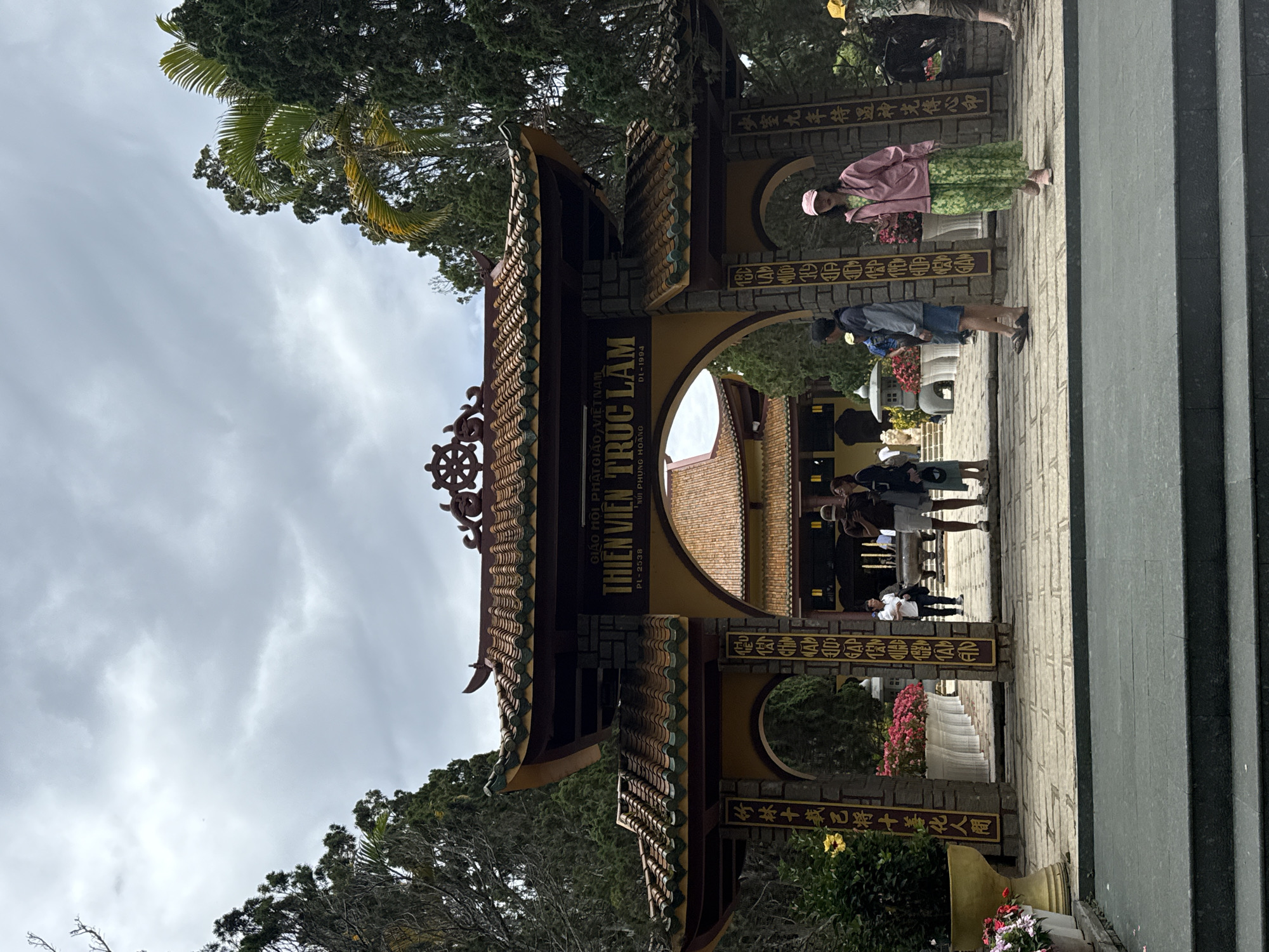 The image shows the entrance to Thien Vien Truc Lam Monastery in Da Lat, Vietnam. The elaborate gate features traditional Vietnamese architecture with layered, curved roofing tiles and intricate carvings. Text in Vietnamese and Chinese characters is inscribed on the gate. People are walking through the gate, including a woman in a pink jacket and green dress. The sky is overcast. Trees surround the entrance. A stone pathway and steps lead to the gate.