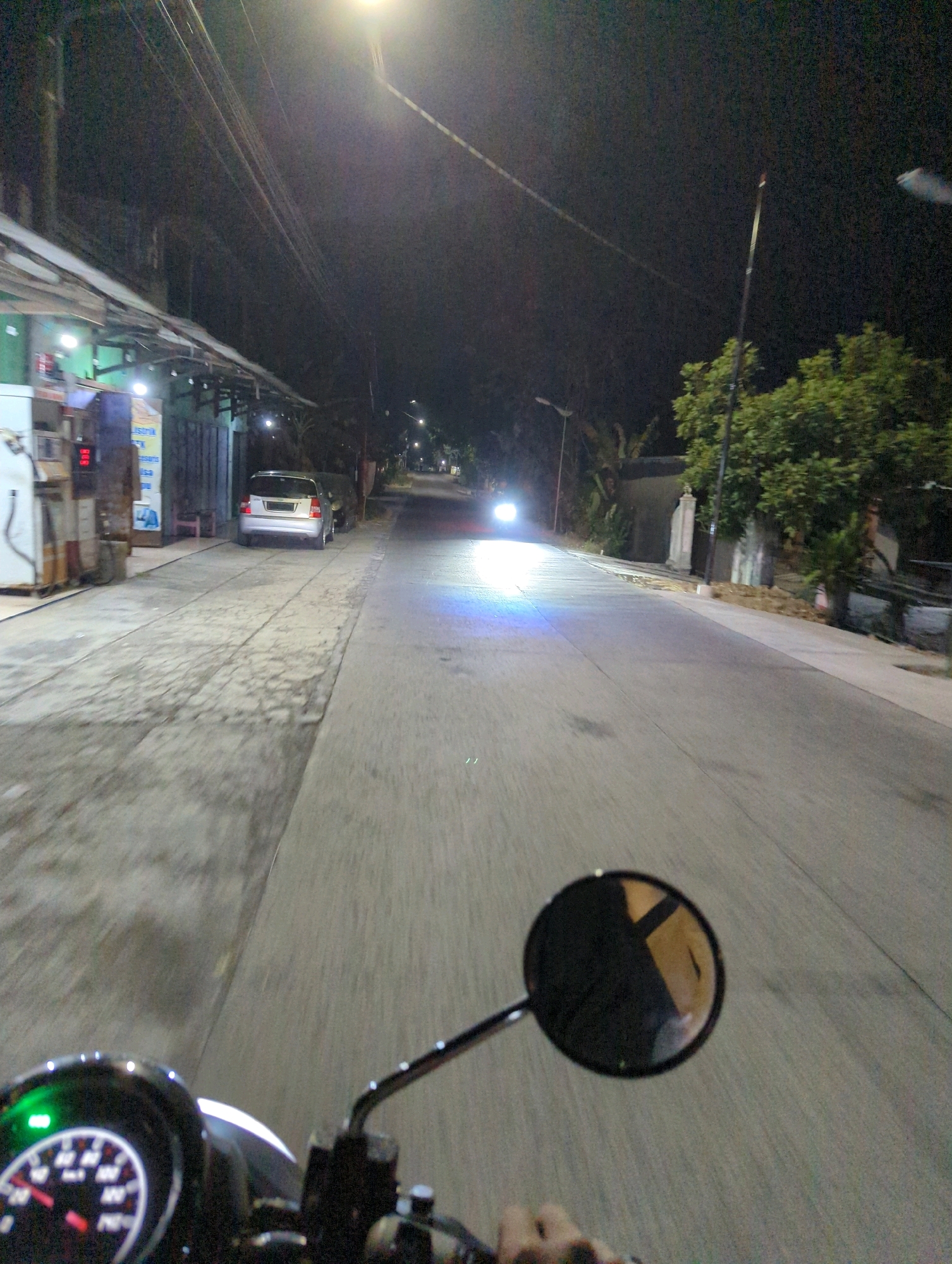 The image depicts a night-time view from the perspective of someone riding a motorcycle in Kacangan, Indonesia. The scene is illuminated by streetlights and the headlights of approaching vehicles. On the left side of the road, there is a building with a green facade, possibly a shop or a gas station, with visible signage. A silver car is parked on the side of the road, facing away from the viewer. The road surface appears to be asphalt, with textured paving on the left side. Overhead, a tangle of electrical wires is visible against the dark sky. Further down the road, a motorcycle's bright headlights are visible, suggesting another vehicle is approaching. The motorcycle the photo is taken from has a visible speedometer with a green indicator light illuminated. A circular rearview mirror on the right side of the handlebars reflects the surroundings. The overall impression is of a typical street scene in a town or city at night.