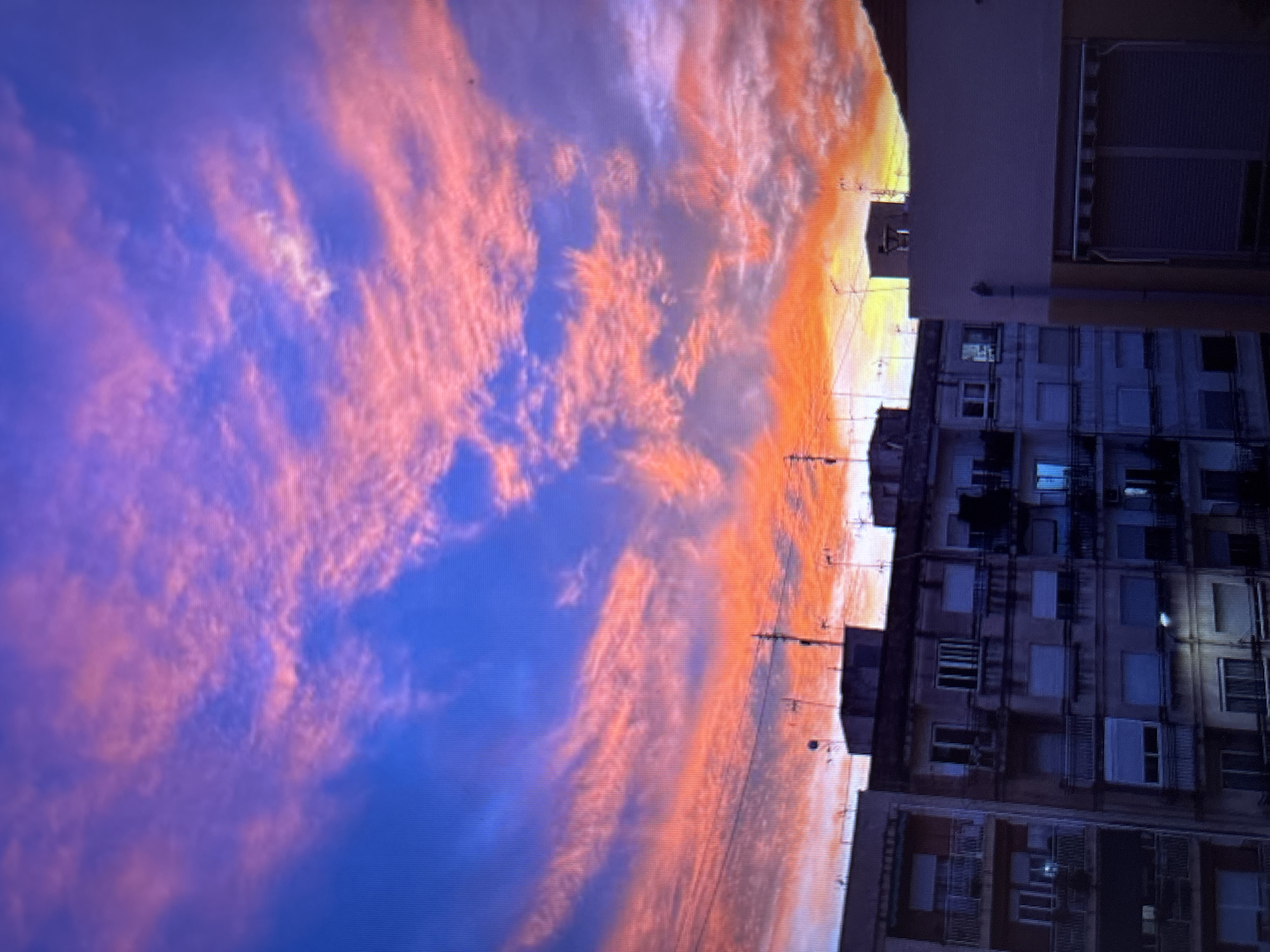 The image shows a city skyline at dusk, with buildings silhouetted against a vibrant sky. The sky is a dramatic mix of deep blues, purples, and fiery oranges and pinks, indicating a sunset or sunrise. Wispy clouds are illuminated by the setting or rising sun, creating streaks of color across the sky. In the foreground, several multi-story apartment buildings are visible. The architecture appears to be typical of a European city, with balconies and numerous windows. The buildings are dark, likely due to the low light conditions of dusk. Antennas and satellite dishes are visible on the rooftops of some buildings. The overall impression is one of urban life under a spectacular natural display in the sky. The scene is devoid of any people or overt human activity. The location is specified as Sofia, Bulgaria, implying this urban landscape is within that city.