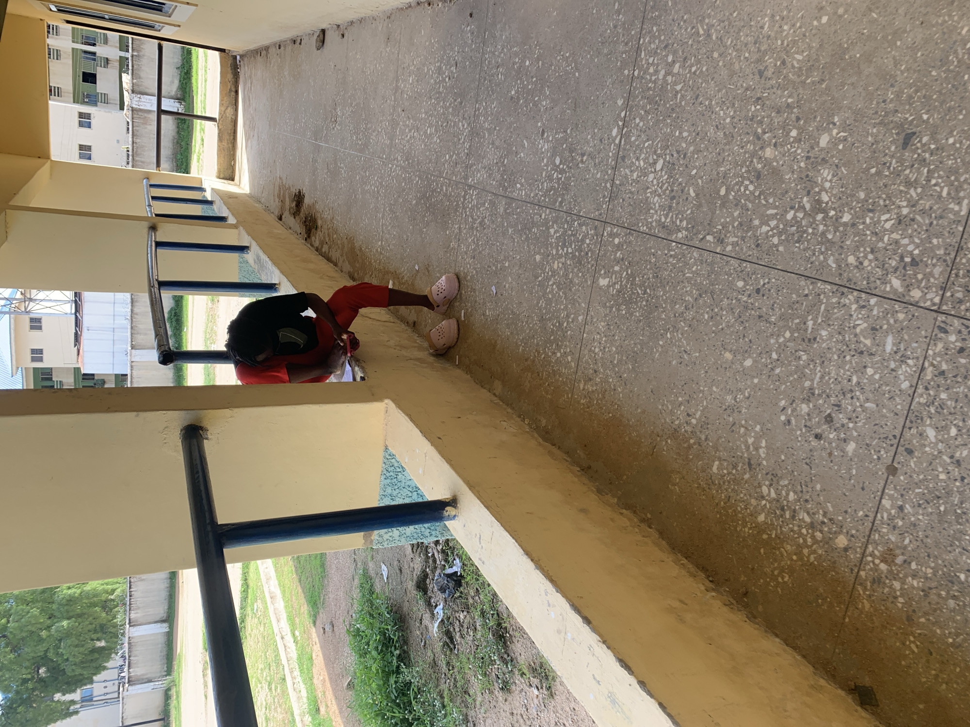 A young person, with dark curly hair, is seated on a low, yellow-painted concrete ledge. They are wearing a red shirt and red pants, with light pink clogs on their feet. The person is looking down at a piece of paper they are holding, appearing to write or read. They are positioned in a covered walkway with a light yellow wall to their right and a blue metal railing to their left. Beyond the railing, there is a grassy area with some scattered litter and a concrete wall with barbed wire on top. In the background, there are multi-story buildings with visible windows. The floor of the walkway is made of grey concrete tiles with speckled aggregate. The scene is outdoors in what appears to be an academic or institutional setting within Jalingo, Nigeria.