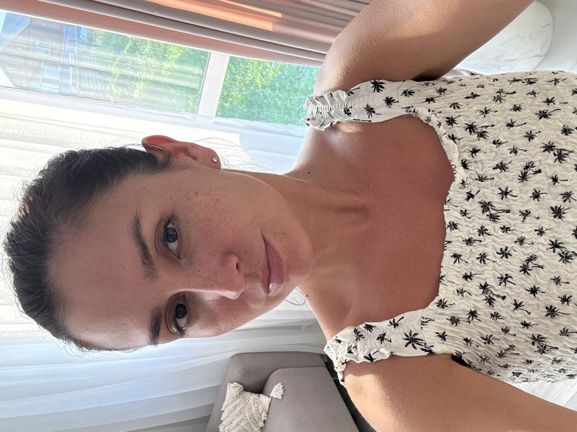 The image shows a woman with dark hair pulled back, wearing a white top with a black palm tree print. She has freckles on her face and is wearing a small diamond stud earring in her left ear. The background features sheer white curtains and a window with a view of a building and greenery. A grey sofa with a textured throw pillow is visible to the left. The lighting suggests it is daytime, possibly indoors with natural light filtering through the window. The location is noted as Nizhnyarkeyevo, Russia.