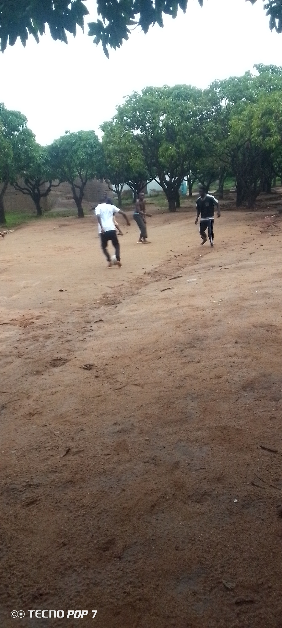 In an outdoor, dusty, rural setting with trees in the background, three young males are engaged in a soccer game. The ground is dry and sandy. One individual, wearing a white t-shirt and dark pants, is in the process of kicking a white soccer ball. Another male, dressed in dark clothing, is facing away from the viewer and appears to be running. A third male, wearing a black and white striped shirt and dark pants, is also running, facing towards the left of the frame. The sky above is overcast.