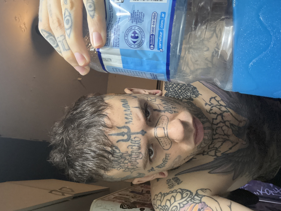 A heavily tattooed individual holds a blue-labeled water bottle. Facial tattoos include "MAMMA," a bandage, and other symbols. Tattoos also cover the hands, neck, and chest, including a wing-like design. The water bottle has text and branding from Carrefour, including a phone number and website. The environment appears to be an interior space, potentially a bedroom, with a corner of a cabinet visible.