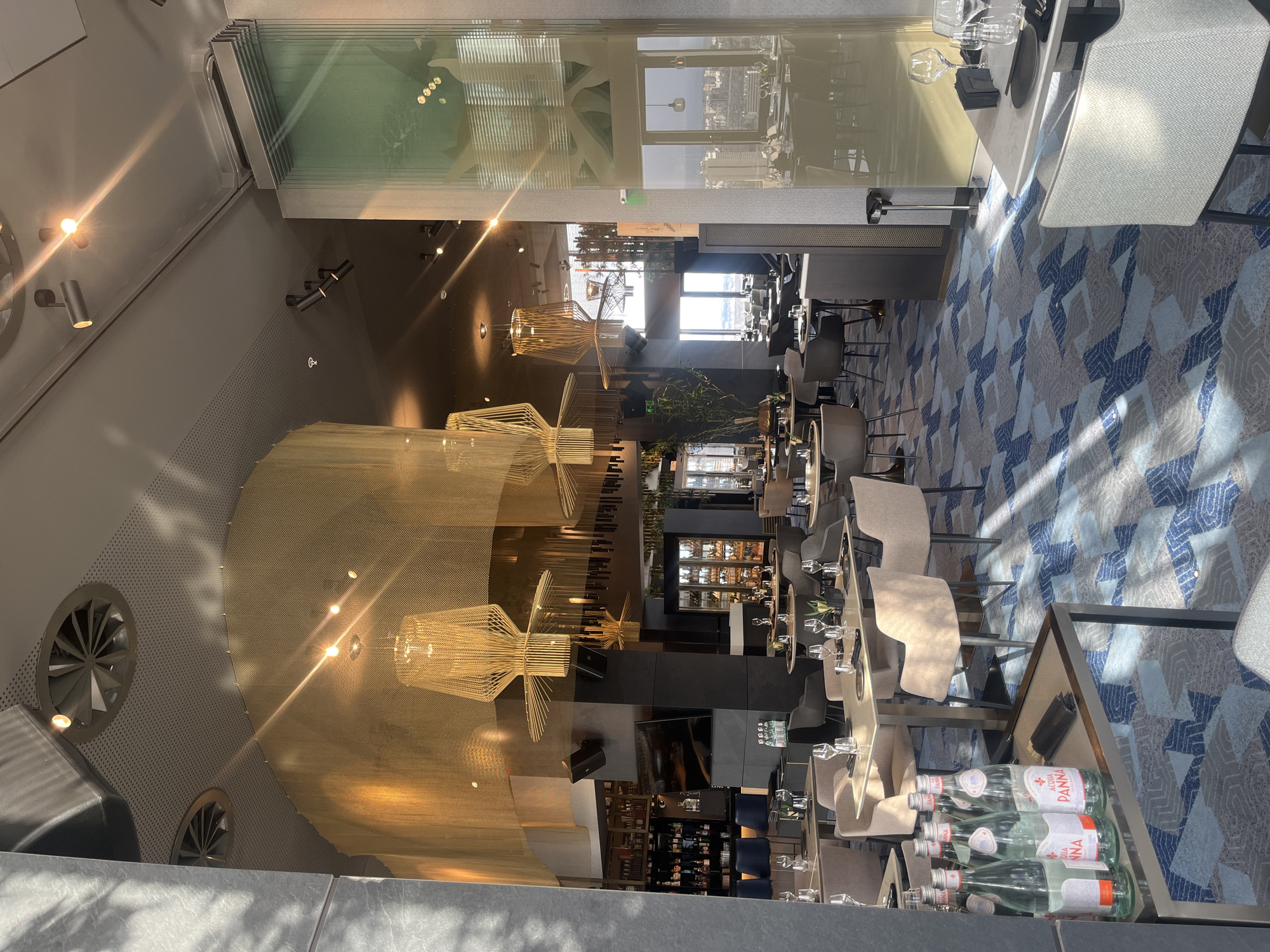 The image depicts the interior of a modern restaurant, likely located in the city of Sofia, Bulgaria. The perspective appears to be from an elevated position. The restaurant features a large, curved wall with decorative mesh-like covering and several geometric light fixtures suspended from the ceiling. The dining area is furnished with tables set for service, including glassware and utensils. Chairs are upholstered in neutral tones. A bar area with seating is visible in the background. A floor-to-ceiling glass enclosure separates the space. There are bottles of Acqua Panna at the bottom of the image. The environment is well-lit, with a mix of natural light and artificial lighting. The floor has a patterned carpet in shades of blue, grey, and white. No people are clearly visible, suggesting the photograph was taken when the restaurant was not yet fully occupied or after closing hours.