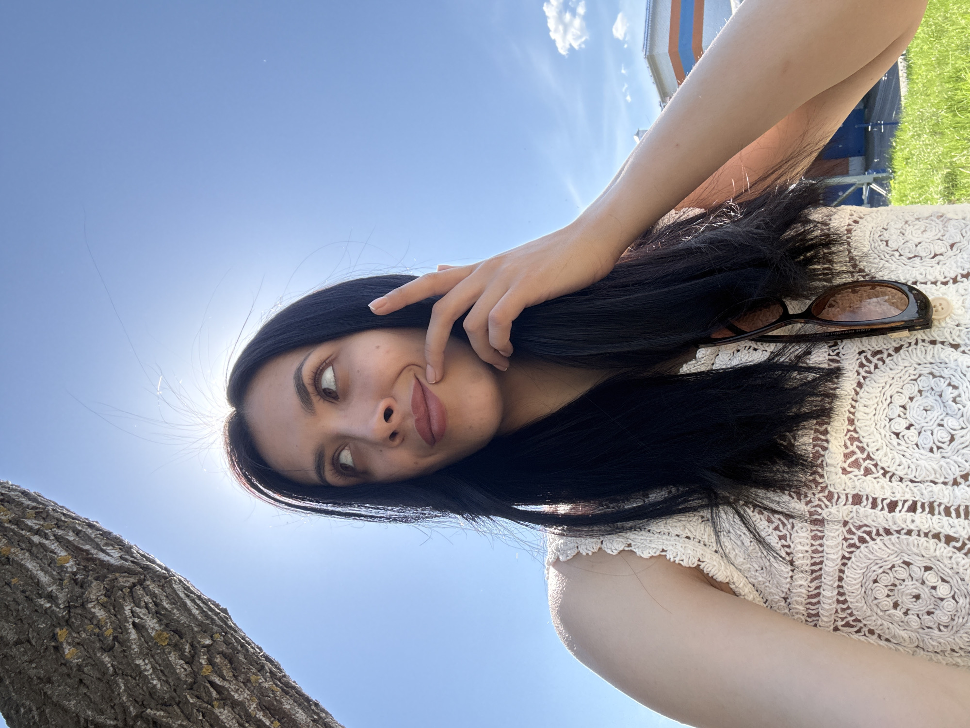A young woman with long dark hair is posing in front of a bright blue sky, likely in an outdoor setting. She is wearing a white, crochet-style top and has a pair of sunglasses perched on her hair. She is holding her hand up to her face and looking off to the side with a playful expression. A tree trunk is visible in the lower left corner. Based on the image, determining the precise location within Votkinsk, Russia is not possible. The setting appears to be a park or outdoor space with trees and open sky. There is a building with blue and orange colors visible at the back right.