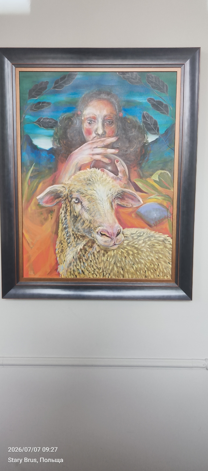 The image displays a framed painting featuring a woman with dark, curly hair and fair skin, her face partially obscured by her hands, which are raised to her mouth. She is depicted with reddish blush on her cheeks and red accents in her hair. In the foreground, a sheep with light-colored wool and pink accents on its nose and ears is shown. The background is a blend of vibrant colors, including blues, oranges, and greens, with stylized leaves framing the top of the painting. The artwork is signed in the lower right corner. The painting is hanging on a light-colored wall, and a date and location stamp in the lower left corner reads "2026/07/07 09:27 Stary Brus, Польща". This indicates the image was taken on July 7, 2026, at 09:27 AM in Stary Brus, Poland. Based on the presence of the text "Stary Brus, Польща" and the fact that the painting is displayed in a location associated with Poland, it is reasonable to infer that the painting is located in Stary Brus, Poland.