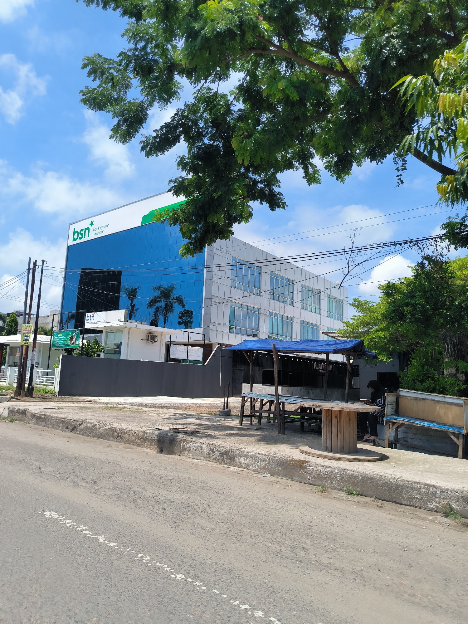 The image captures a street view in Lhokseumawe, Indonesia, focusing on a modern, glass-fronted building. The building features a prominent "BSN bank syariah nasional" logo. In front of the building, there's a shaded outdoor seating area with wooden tables and benches, and a person is seated at one of the tables, looking at their phone. The street in the foreground is paved with asphalt and has a concrete curb. Overhead, there are power lines and lush green trees with foliage obscuring parts of the sky and building. The sky is a clear blue with scattered white clouds.