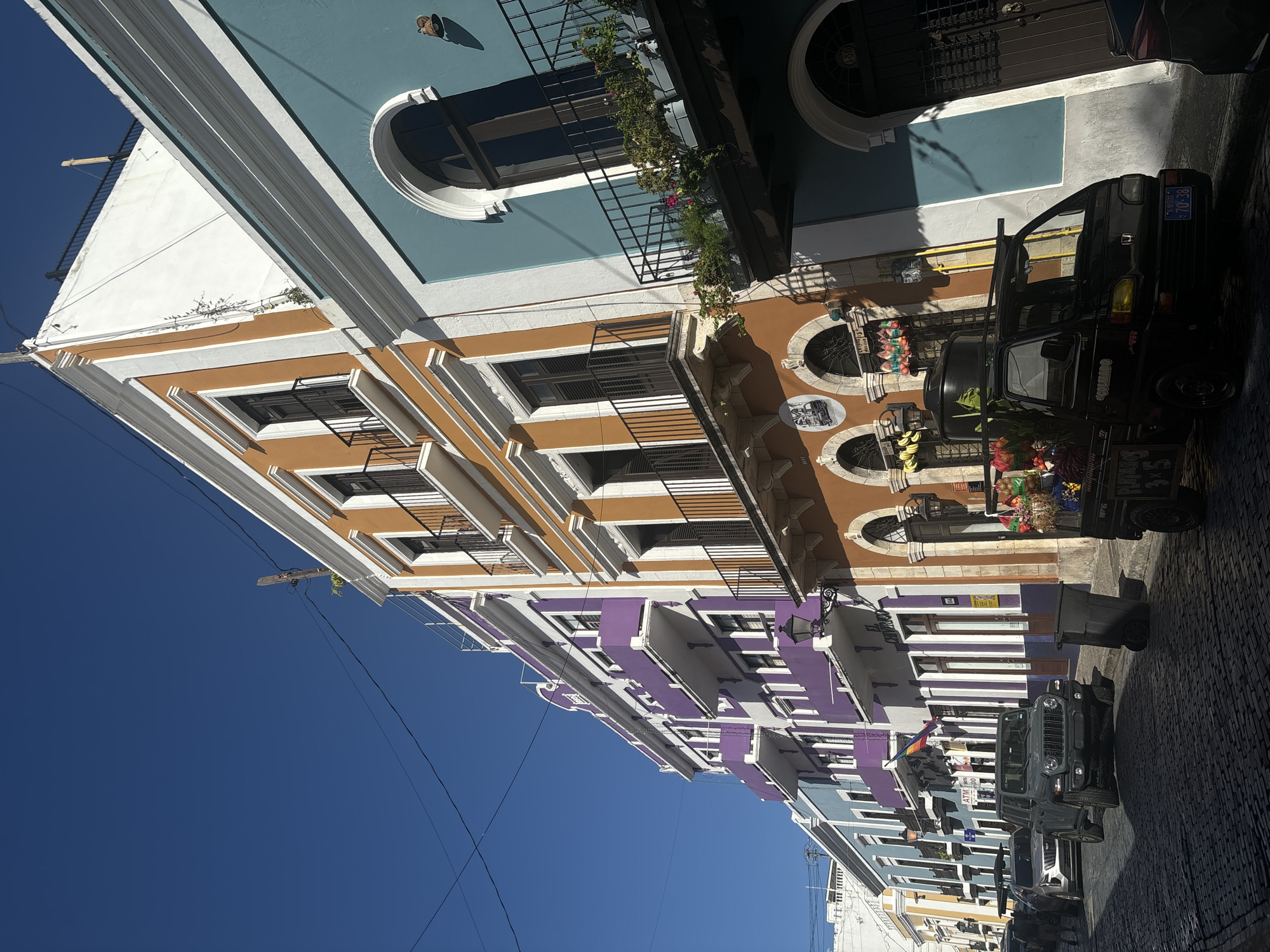 The image depicts a street scene in Old San Juan, Puerto Rico, characterized by colorful colonial-era buildings lining a cobblestone street. The buildings feature vibrant facades in shades of orange, purple, and turquoise, with white trim and wrought-iron balconies. Several cars are parked or driving on the street, including a gray Jeep and a black mini-truck laden with flowers. The sky is a clear, deep blue, suggesting a sunny day. The architecture, the street surface, and the style of the vehicles all point to a historic urban setting in a tropical climate. There are no visible people in the immediate foreground. The overall impression is one of a lively, picturesque street in a well-preserved historic district.