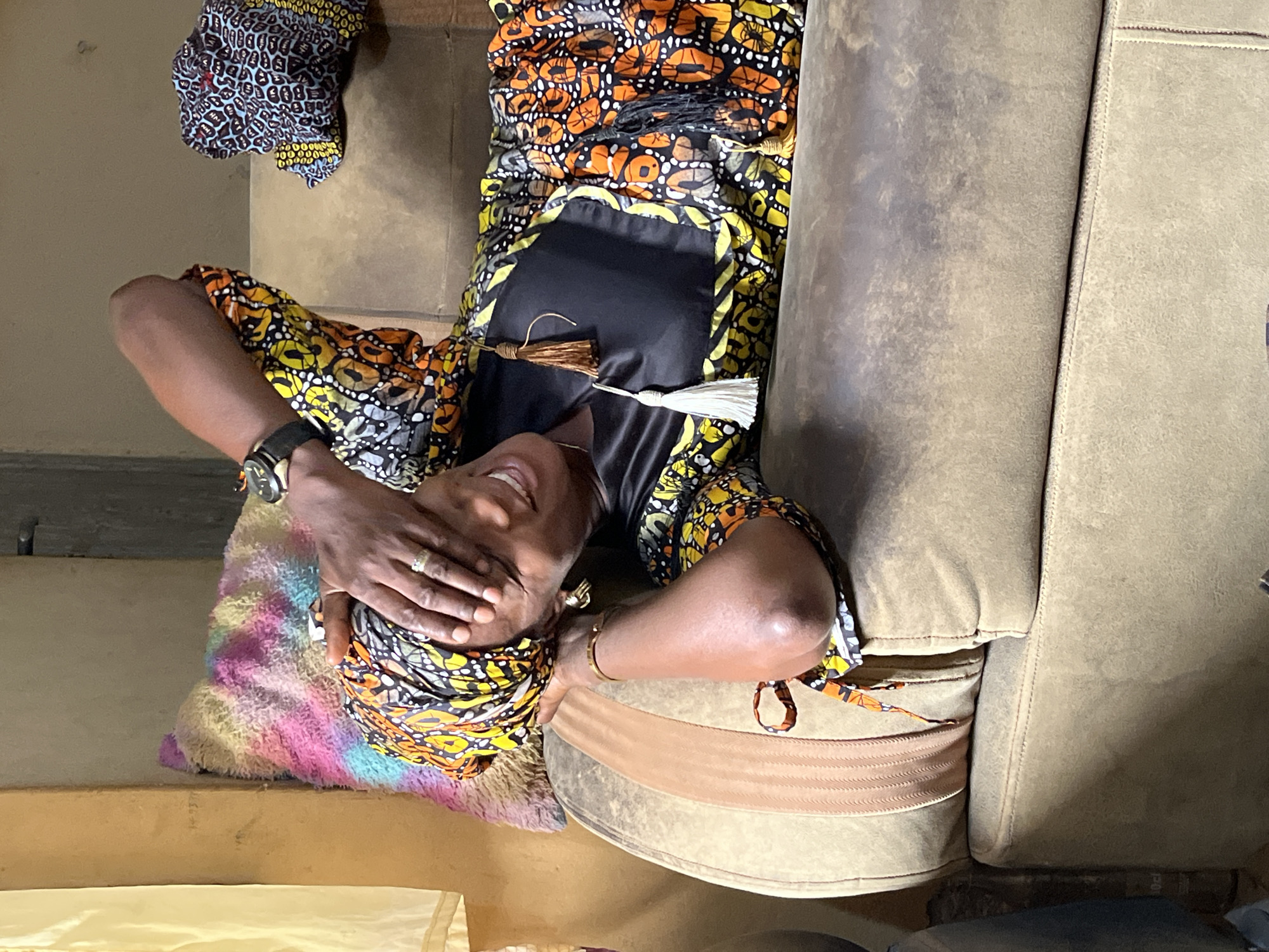A woman is lying down on a light-brown colored couch with a colorful pillow supporting her head. She is wearing a black and colorful print dress and a matching head wrap. Her left hand is covering her eyes. She has on a watch and jewelry. The location is Madalla, Nigeria.
