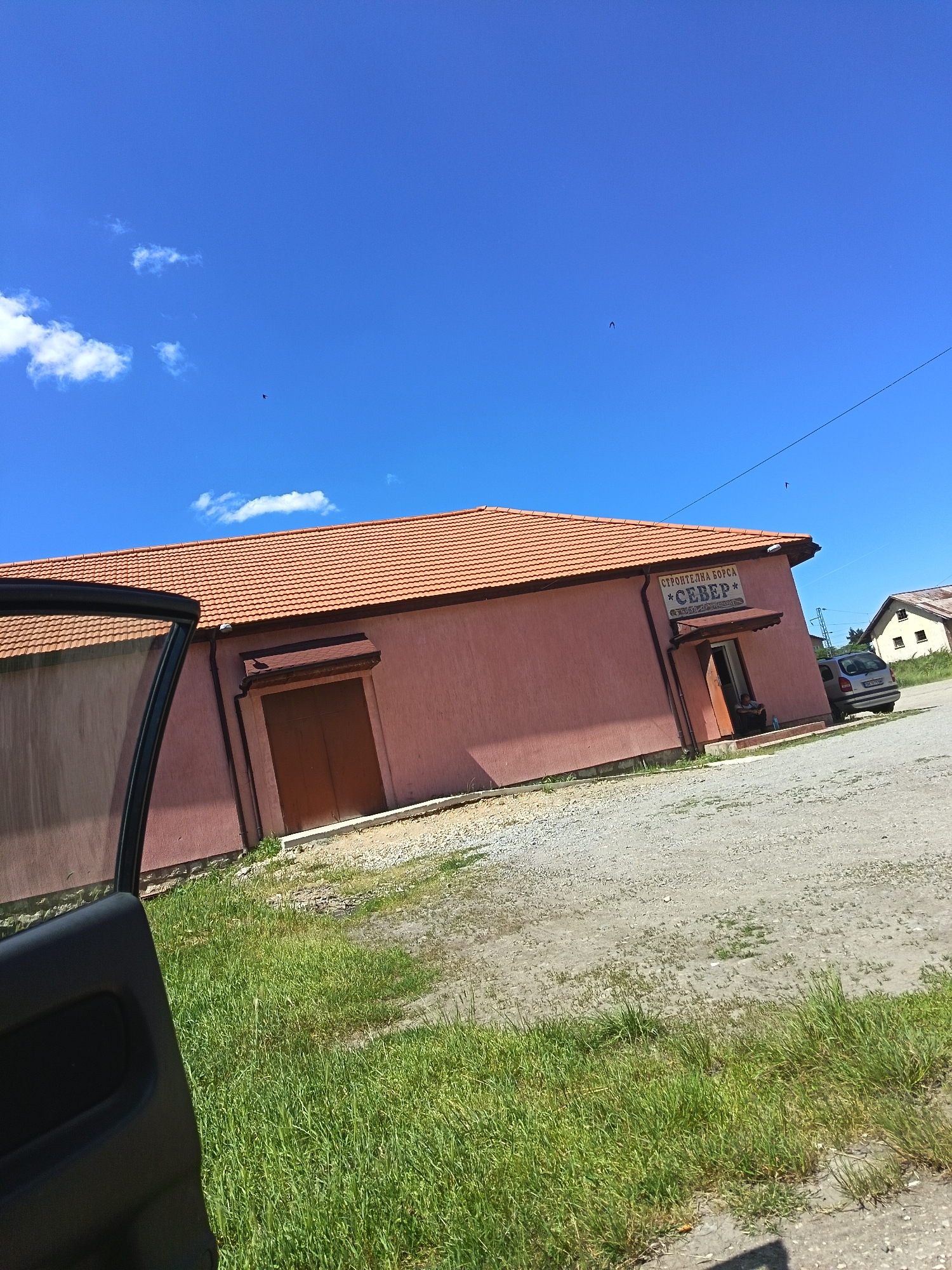 The image shows a one-story building with a terracotta tile roof in Drenovets, Bulgaria. The building has a salmon-colored exterior with brown doors and an awning above one of the entrances. Above the entrance, there is a sign that reads "СТРОИТЕЛНА БОРСА СЕВЕР." Adjacent to the building is a gravel parking area and a patch of green grass. A car is parked near the building. The sky is blue with a few scattered clouds. A portion of a car door is visible in the foreground on the left.
