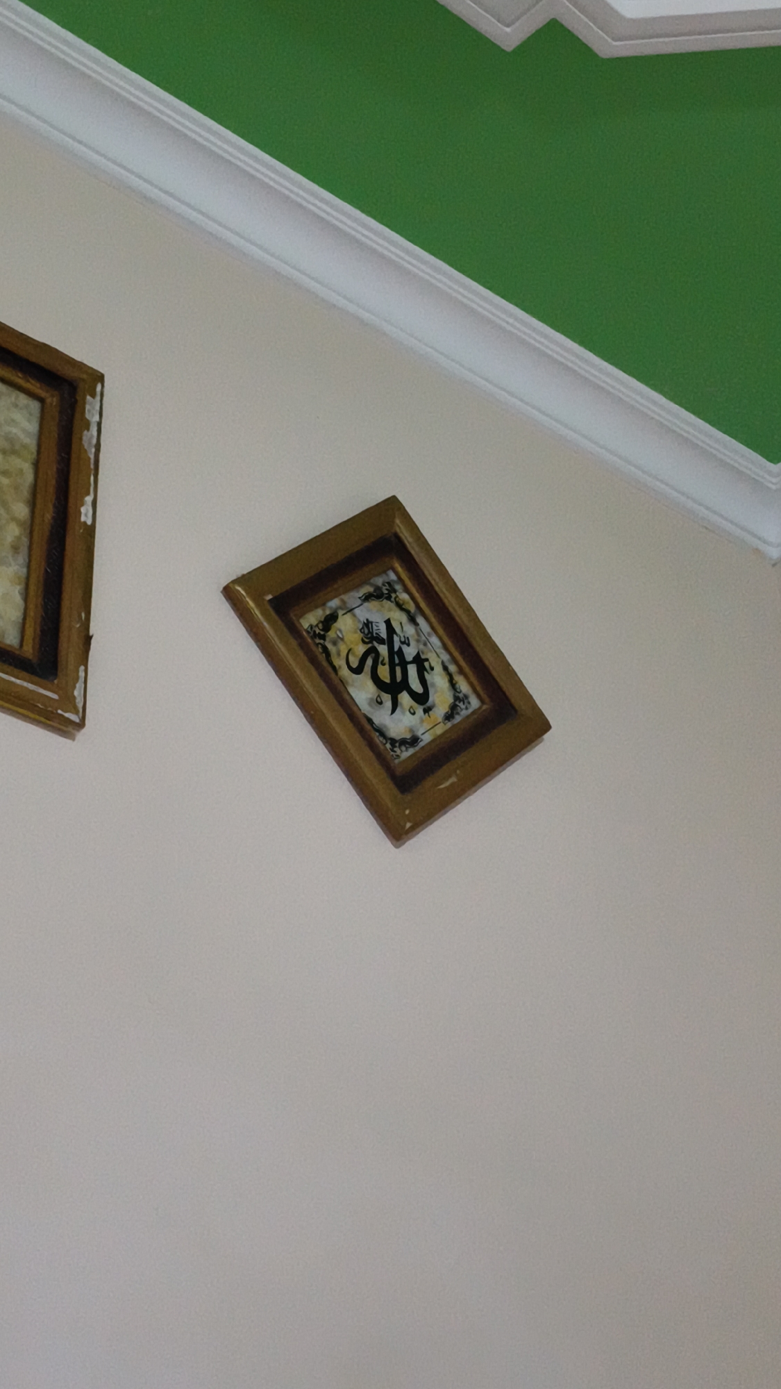A framed Islamic calligraphy is displayed on a light-colored wall. The calligraphy appears to be in black Arabic script on a patterned background within the frame. The frame itself is made of wood and has a golden hue. To the left of this frame, another framed artwork is partially visible, also with a wooden frame. Above the wall, a section of a bright green ceiling is visible, separated from the wall by white molding. The scene is captured from a low angle, looking upwards. The location is in Kerep Kidul, Indonesia.