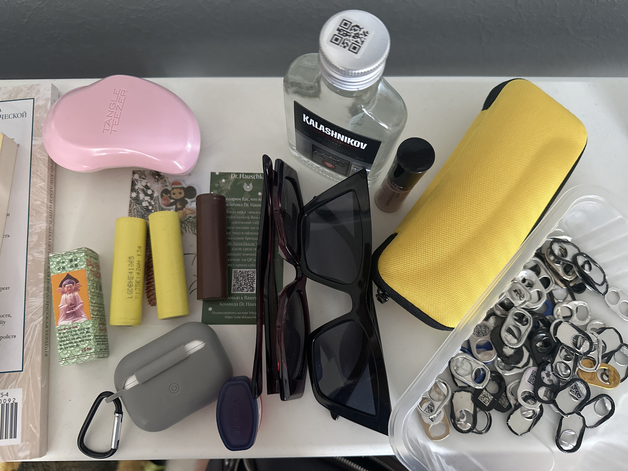 The image depicts a collection of personal items arranged on a white surface, likely a shelf or tabletop. In the background, a book or magazine with visible text and what appears to be a barcode is present.
Prominent objects include:
- A light pink, ergonomically shaped hairbrush with the brand name "TANGLE TEEZER" embossed on it.
- A grey earbud case with a carabiner clip attached.
- Two yellow cylindrical objects, resembling batteries or power banks, with text visible on them.
- A small, rectangular brown object.
- A small blue container with a red lid, possibly containing a cosmetic product.
- A pair of dark-rimmed sunglasses.
- Another pair of black, oversized sunglasses.
- A clear glass bottle with a silver cap containing a clear liquid, labeled "KALASHNIKOV". A QR code is affixed to the cap.
- A small dark bottle with a black cap, possibly an essential oil or perfume.
- A yellow zippered case, possibly for glasses or a small electronic device.
- A clear plastic container filled with numerous metal pull tabs, some of which have QR codes printed on them.
The overall arrangement suggests a casual, personal setting. There are no people visible in the image, nor any explicit indication of the city of Nevinnomyssk, Russia. The lighting is neutral, and the focus is on the objects themselves. The scene is static, with no observable actions or interactions taking place. The surfaces appear clean and organized.