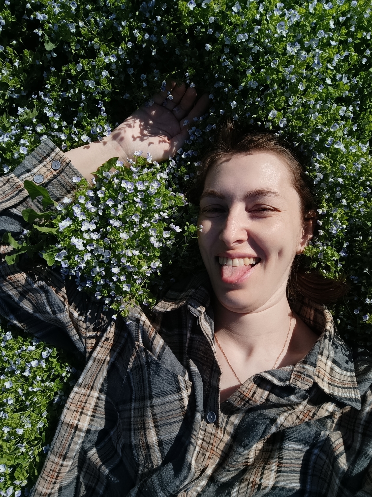 A young woman lies on her back in a field of small, light-blue flowers. She is wearing a plaid shirt and has her arm raised above her head. Her eyes are partially closed, and she is sticking her tongue out with a playful expression. The scene appears to be outdoors, and the abundance of flowers suggests it could be a field or park. It is daytime and sunny, indicated by the shadows on her hand. It is impossible to verify the location as the city of Chaikovsky, Russia, from the image alone.