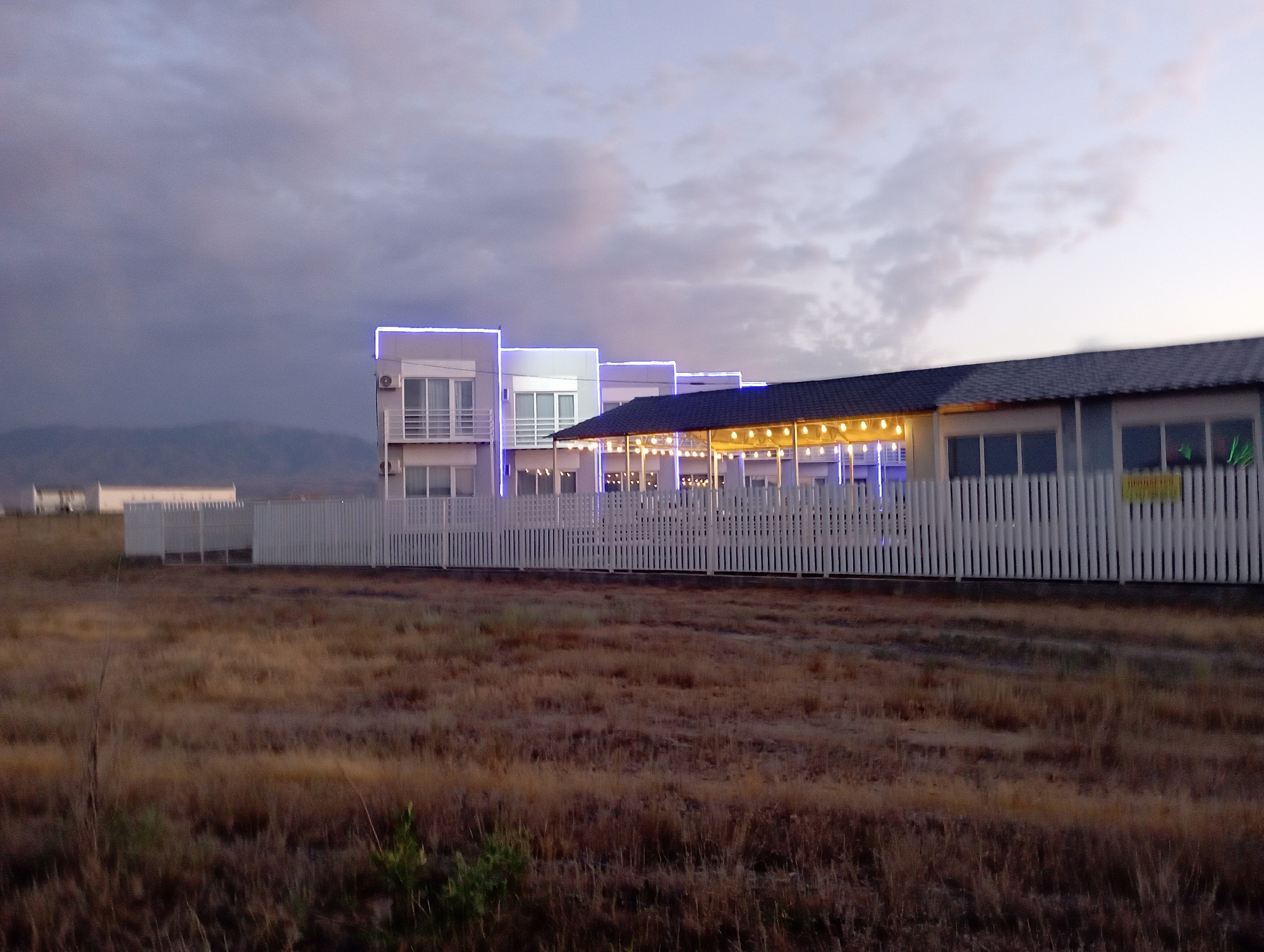 In an outdoor scene, a modern, multi-story building complex with white facades is illuminated by blue and yellow string lights, suggesting evening or dusk. The complex features multiple levels with balconies and large windows, and a long, white picket fence separates the property from an open field of dry, brown grass. In the background, distant hills are visible under a dusky, cloudy sky. The scene is devoid of any people or visible activity, presenting a static and empty environment.