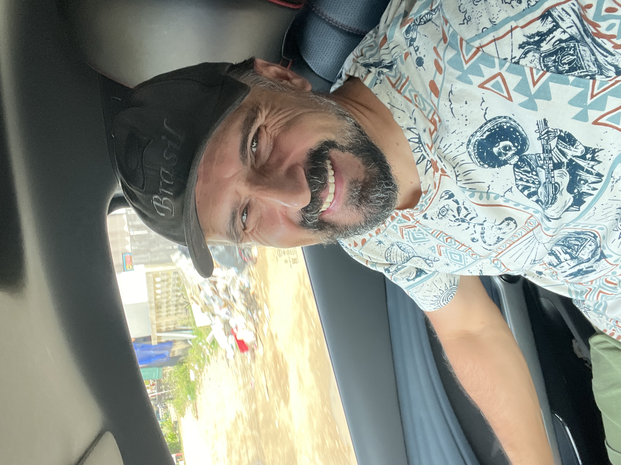 A man wearing a black baseball cap that says "Brasil" and a white t-shirt with a patterned print is sitting in the driver's seat of a car. He has a gray beard and is smiling. The car's interior is visible, with a gray headliner and dark seats. Through the window, a dirt road is visible, with a pile of trash and some buildings in the background. The image appears to be taken in Baan Plai Laem, Thailand.