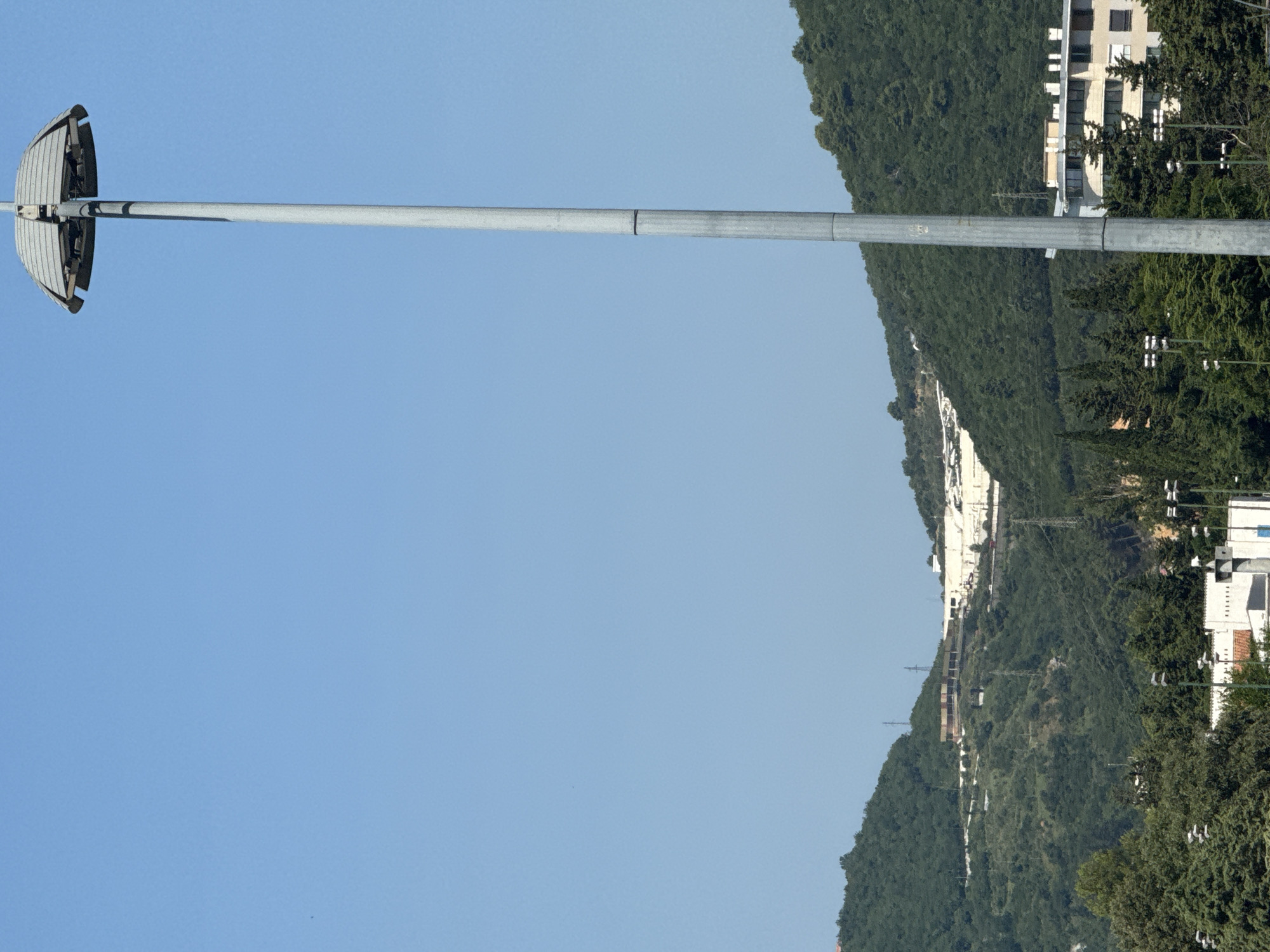 The image shows a low-angle view of a large, parabolic reflector mounted on a tall metal pole against a clear blue sky. The reflector is made of horizontal slats and is positioned to the left of the frame. A long, thin metal pole extends horizontally from the pole supporting the reflector, disappearing to the right.
In the background, a lush, green, densely forested hillside rises steeply. Along the hillside, several white buildings with multiple windows are visible, interspersed with trees. Power lines and utility poles are also scattered across the slope. The buildings appear to be multi-story structures. The overall impression is a daytime scene under fair weather conditions, with a view of a mountainous coastal area. The location is identified as Budva, Montenegro. There is no visible text in the image.