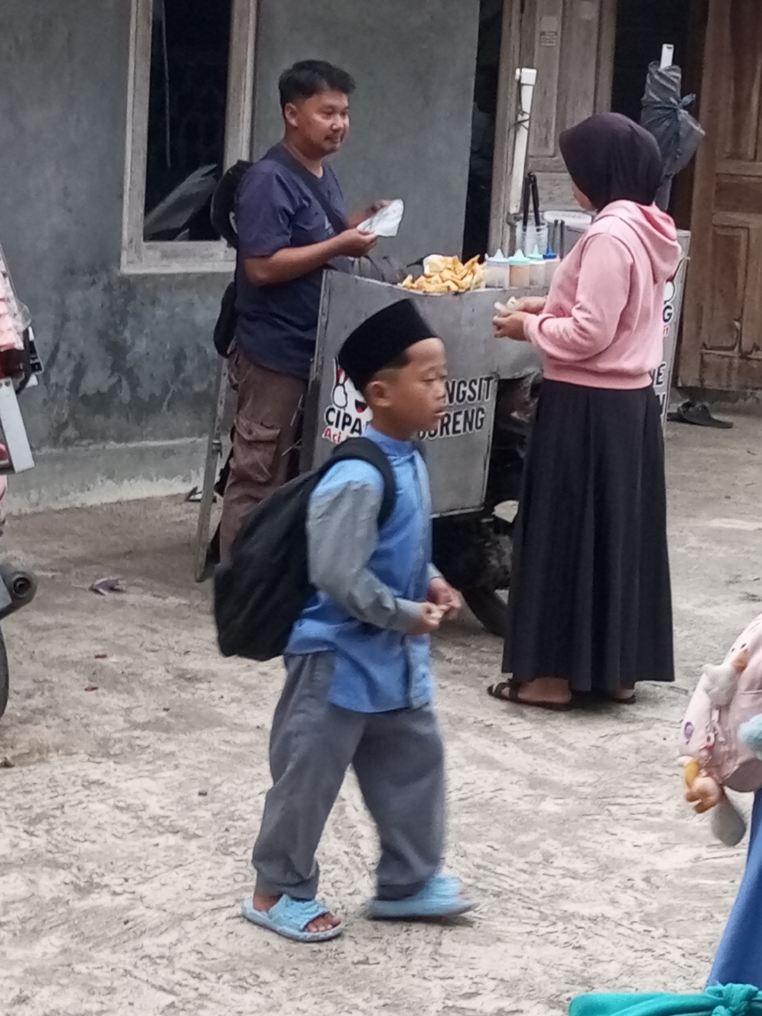 The image shows a street scene in Krasak, Indonesia. In the foreground, a young boy wearing a blue tunic, grey pants, a black skullcap, and blue sandals walks from left to right, carrying a black backpack. Behind him, an adult man in a dark blue t-shirt and brown pants stands near a food cart, holding a white object. To the right of the food cart, a woman in a pink hoodie and a black hijab and skirt stands, holding a white object. The food cart appears to be selling snacks or street food. The ground is paved with rough concrete. The background shows a building with a wooden door and a window.