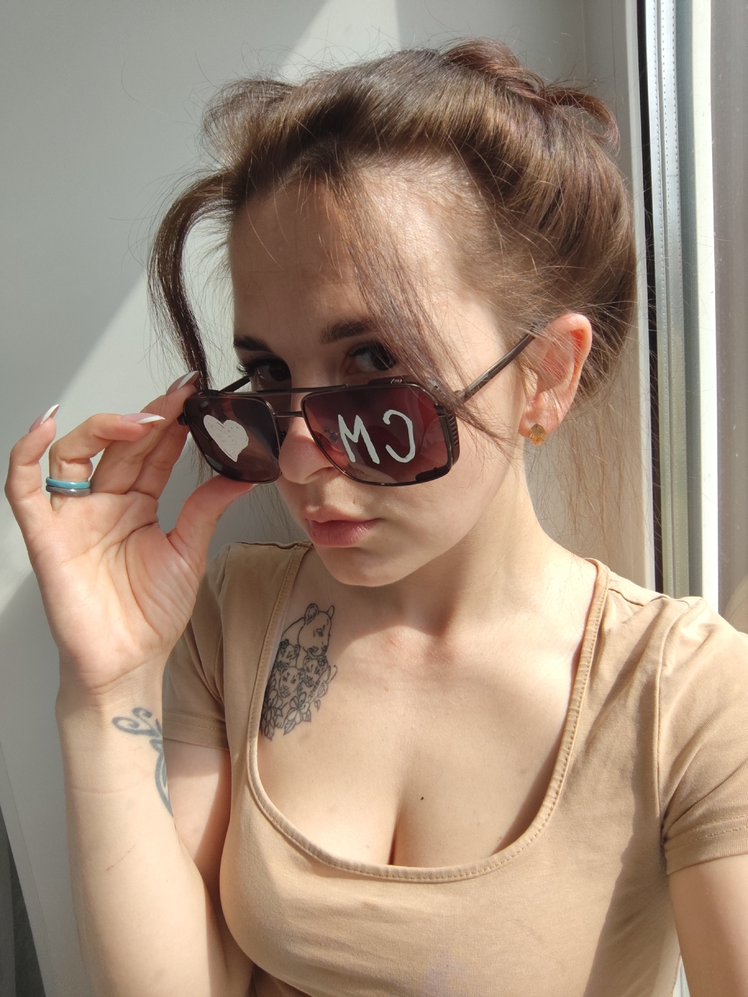 A young woman is depicted in a well-lit, close-up indoor portrait, likely taken in Фоки, Russia. She has light brown hair, pulled back with some loose strands framing her face, and looks directly at the camera with a focused, neutral expression.
She wears a simple light beige t-shirt with a scoop neckline, revealing a tattoo of stylized animal figures on her upper chest/shoulder. Another tattoo with a swirling design is visible on her left forearm. Her left hand, adorned with long, white-tipped nails, holds down the arm of her dark-lensed sunglasses, allowing her to peer over them. On her left ring finger, she wears two rings, one blue and one grey. A small, golden stud earring is visible in her right ear.
The sunglasses have white markings on their lenses: a heart and the letters "M♥O" are visible on the left lens (from her perspective), while "CM" appears on the right lens, reversed as seen by the viewer.
The background consists of a plain, light-colored wall with strong, angular shadows cast by a window frame, which is visible on the right side of the image. The bright, natural light indicates a sunny day, likely morning or afternoon, streaming into the room.
