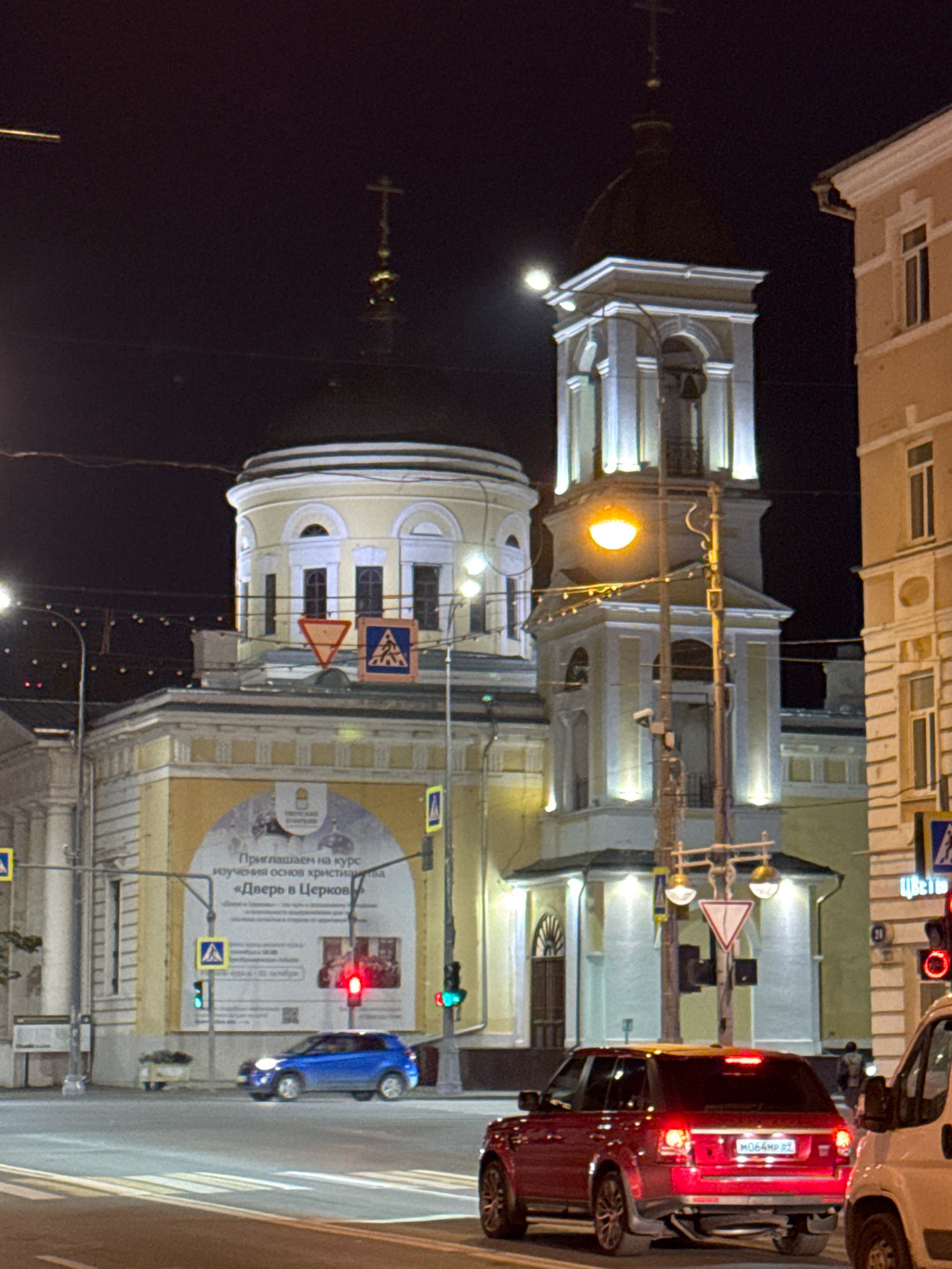 The image depicts a nighttime view of a street scene in Tver, Russia, featuring a church with a prominent bell tower and a light-yellow domed structure. The architecture appears to be traditional Russian Orthodox. The buildings are illuminated by streetlights and architectural lighting, creating a dramatic effect against the dark sky.
In the foreground, a red Range Rover is parked, facing away from the viewer, with its brake lights illuminated. To its right, a portion of a white van is visible. A blue hatchback car is driving away from the viewer on the street. Traffic lights are visible, one showing red and another green.
Several road signs are present, including a yield sign and pedestrian crossing signs. A large banner advertising a course on "The Door to the Church" is displayed on the side of the yellow building. The street is paved, and pedestrian crossings are marked. The overall impression is of a well-lit urban environment at night, with religious and civic structures dominating the scene. There are no visible people in clear focus.
