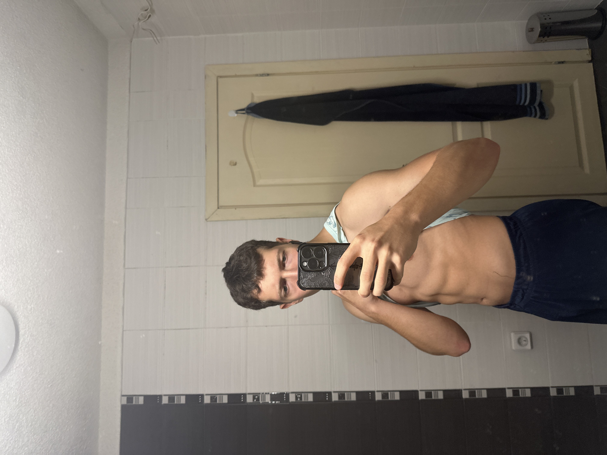 In a bathroom, a young man with short, dark hair takes a mirror selfie. He is wearing dark blue sweatpants and a light blue tank top, which he is lifting to expose his abs. He holds a black smartphone in his right hand, angled to capture his reflection. The bathroom walls are tiled with white and dark grey rectangular tiles. To the left, a section of dark tiles with a repeating black and white pattern is visible. A light-colored door with a curved top is behind him, with a dark garment hanging on a hook to the right of the door. A silver trash can is located on the floor to the right. An electrical outlet is visible on the tiled wall behind him. A circular ceiling light fixture is present above.