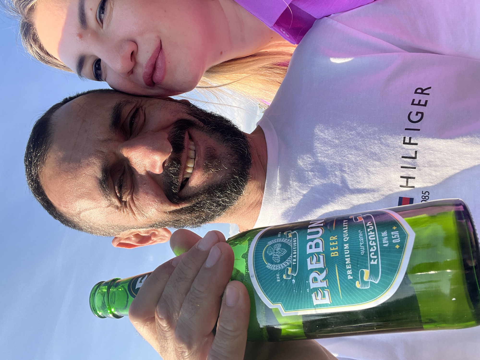 A man and a woman are pictured in a close-up selfie, against a bright blue sky. The man, holding a green bottle of Erebuni beer, is on the left, smiling with his eyes closed and a beard. He is wearing a white t-shirt with the Tommy Hilfiger logo visible. The woman, to his right, has blonde hair and is leaning her head towards his. She is wearing a bright pink or purple top. The beer bottle has a green label with text in both English and Armenian script. The sun is shining brightly, casting light on their faces. The background is a clear blue sky, suggesting an outdoor setting. The location is identified as Yanovo-Grushevsky, Russia.