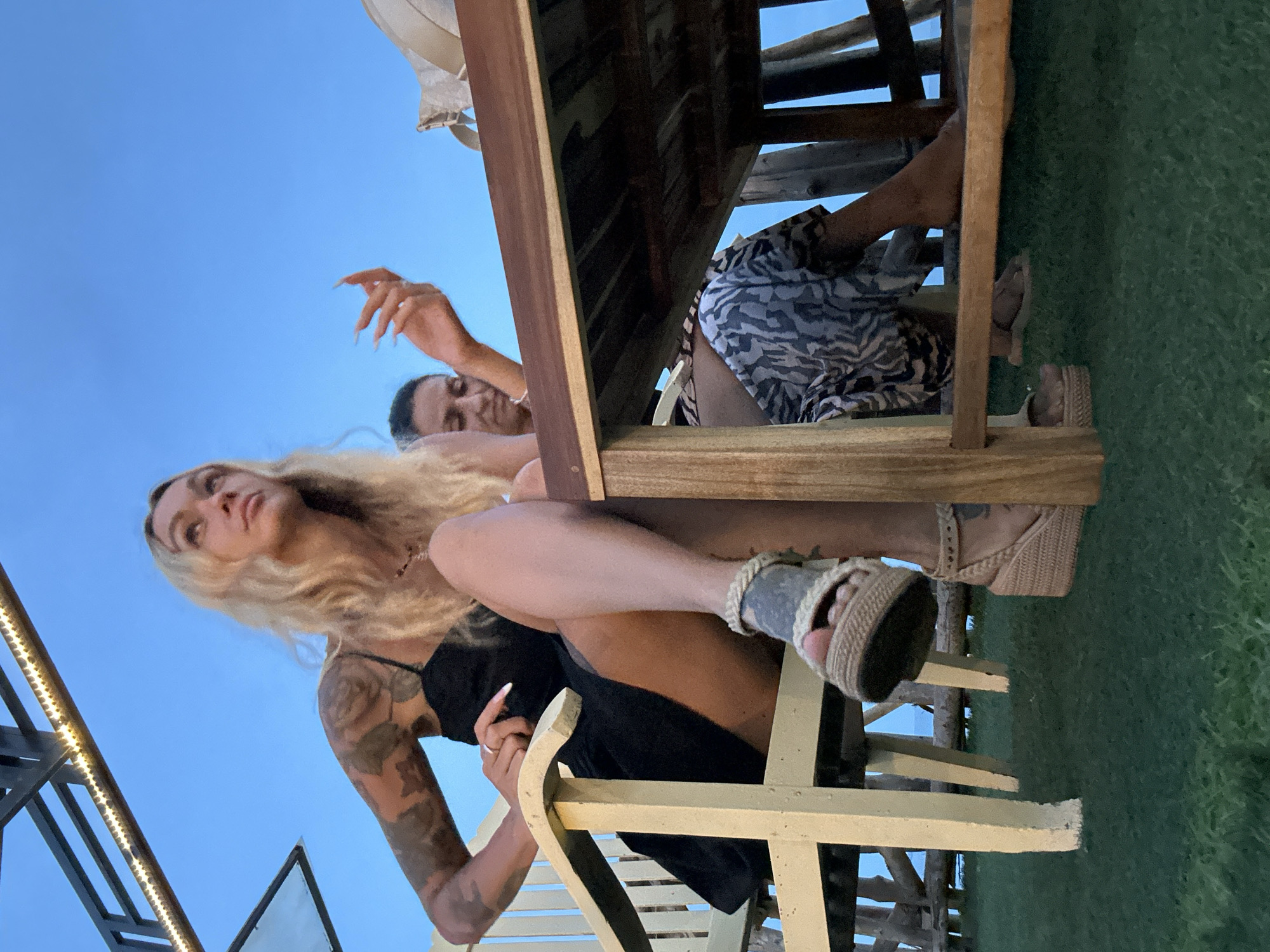 The image shows two women sitting outdoors in what appears to be a restaurant or bar area with a view of the ocean. The woman in the foreground, with blonde hair and tattoos on her arm, is wearing a black dress and platform sandals. She is seated in a light-colored chair, leaning back with one leg crossed over the other. The second woman, with dark hair, is seated behind a wooden table and is wearing a patterned dress. She is looking down with her hand raised. The environment includes a wooden table, chairs, and a green grassy area. The sky is a clear blue, suggesting it is either late afternoon or early evening. The location is identified as Phu Thuy Ward, Vietnam.