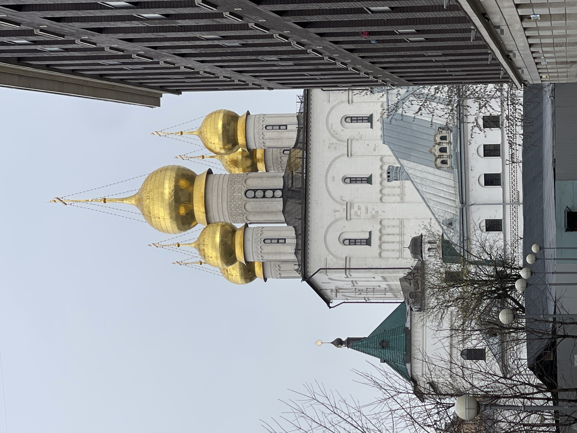 The image depicts a view of the Moscow Kremlin's Cathedral of the Archangel, a prominent Russian Orthodox church in Moscow, Russia. It is characterized by its white stone walls, gilded onion domes, and Renaissance-inspired architectural features. The adjacent modern building contrasts with the historic architecture. The scene likely takes place on a cloudy day.