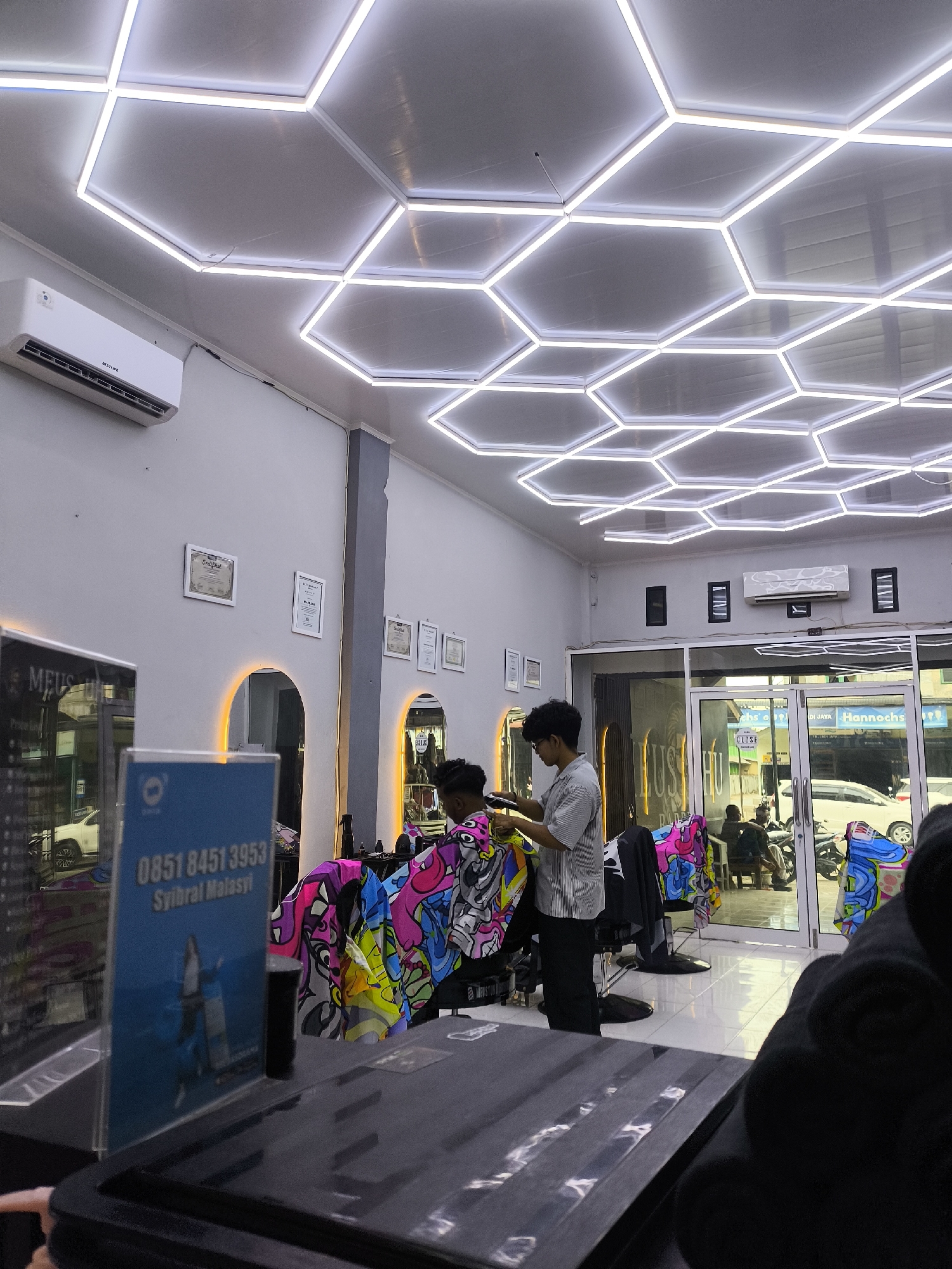 The image depicts the interior of a barber shop located in Miruektaman, Indonesia. The shop features a modern design with a hexagonal LED lighting system on the ceiling and recessed arch lighting behind mirrors. An air conditioning unit is mounted on the wall. In the foreground, a barber is attending to a client, using clippers. The client is seated, wearing a vibrantly patterned cape. To the left, a blue sign displays a phone number "0851 8451 3953" and the name "Syibrall Malasyi". Reflections in a mirror on the left show the exterior of the shop, including a white car. Through the glass doors at the front, other patrons can be seen sitting and interacting, with motorcycles parked outside. The overall atmosphere suggests a contemporary and well-lit establishment.