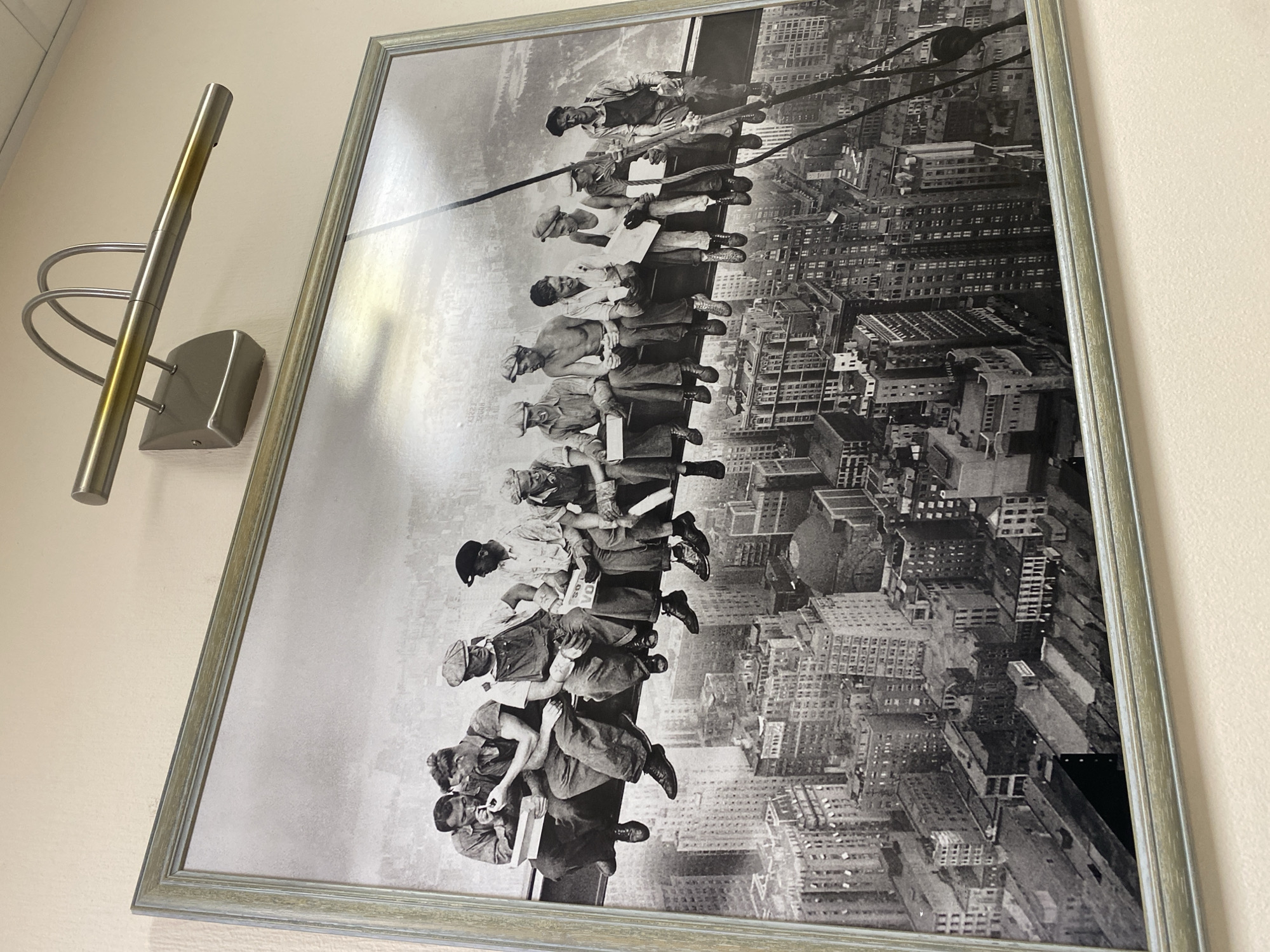 The image shows a framed, black-and-white photograph displayed on a plain, light-colored wall. To the left of the framed picture, a modern, wall-mounted picture light with a brushed metal finish, featuring a horizontal bar and curved supports, is visible.
The photograph depicts the iconic "Lunch atop a Skyscraper," featuring eleven construction workers seated in a row on a steel beam high above a bustling city. The workers, dressed in typical work attire including hats and boots, appear to be on a break, with several smoking cigarettes and others holding food or drink containers. One worker is seen lighting another's cigarette, while others converse or look out at the panoramic cityscape below. A newspaper or piece of paper is held by one worker, and another holds a small sign with the word "VOTE" clearly visible.
The background reveals a dense urban landscape of numerous buildings stretching into the horizon, indicative of a major city during a period of significant construction. The scene is set on a clear, bright day. The workers' demeanor suggests ease and camaraderie despite their perilous perch. A noticeable reflection from a light source is present on the surface of the framed photograph, especially across its upper portion. The frame itself is a light-toned, possibly distressed metallic or wooden design.