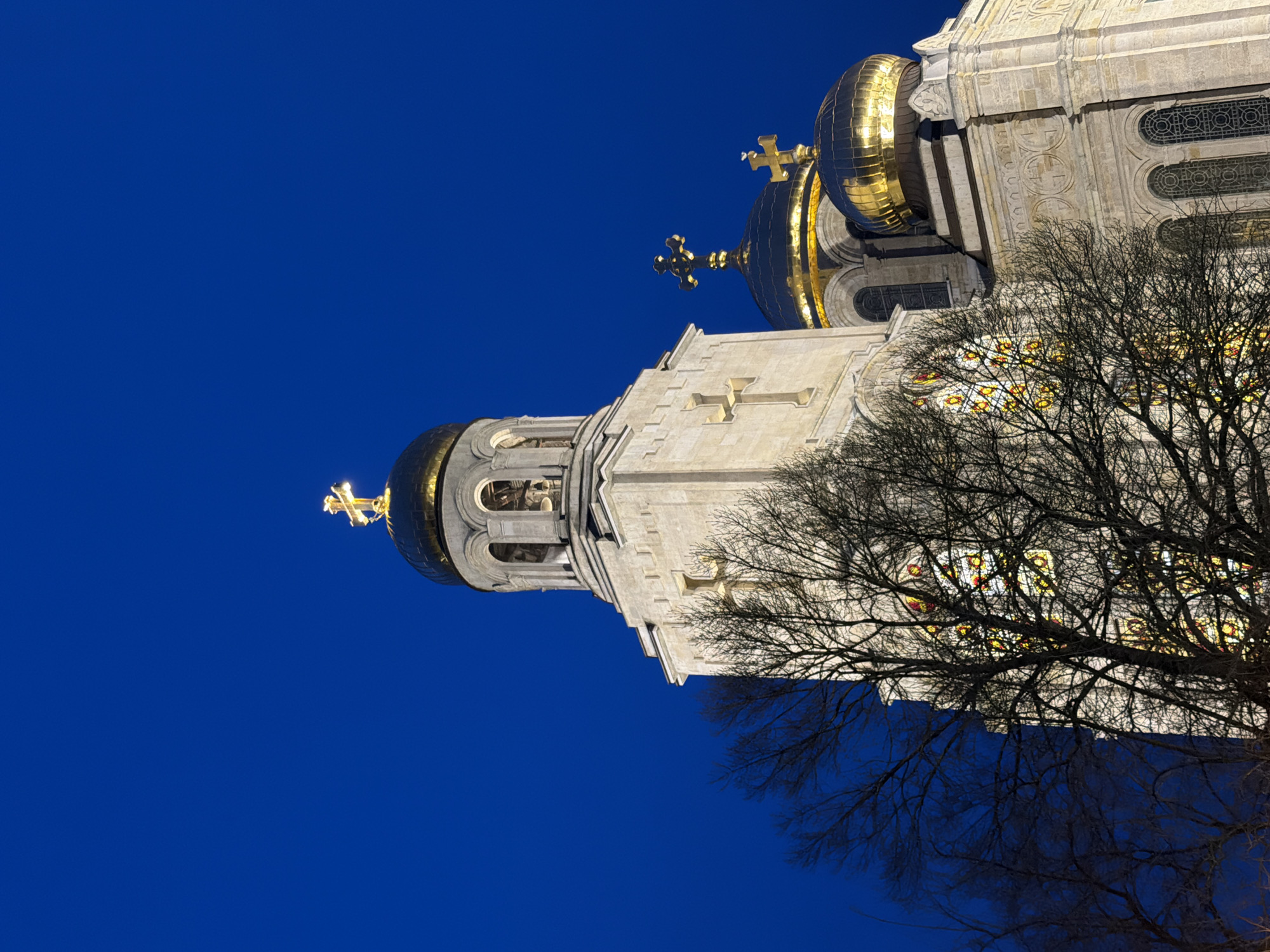 The image shows a low-angle view of a building with distinctive architectural features, set against a clear, deep blue sky. The building appears to be a church or cathedral, characterized by a pale stone facade and golden, onion-shaped domes topped with crosses. A bare tree with intricate branches partially obscures the building's facade, adding depth and texture to the composition. Stained glass windows can be seen through the branches. The scene is likely a view of the Dormition of the Mother of God Cathedral in Varna, Bulgaria.