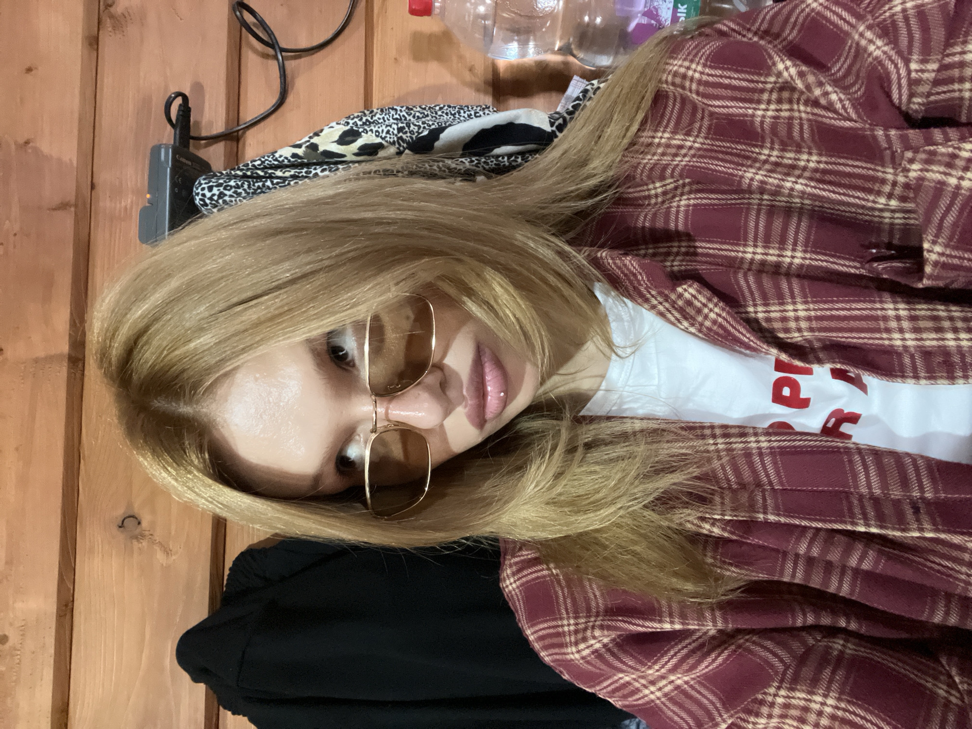 A young woman with blonde hair is looking to her left, wearing oversized sunglasses with gold frames, a white t-shirt with red lettering, and an open maroon and beige plaid shirt. She has a silver lip ring. She is indoors, with a wooden plank wall behind her. To her left, a black garment hangs on a hook. To her right, a leopard-print scarf is draped over a black object, and a partially visible plastic bottle of water with a red cap is also present. The background suggests a rustic or unfinished interior, possibly a cabin or workshop. The lighting is uneven, casting shadows.