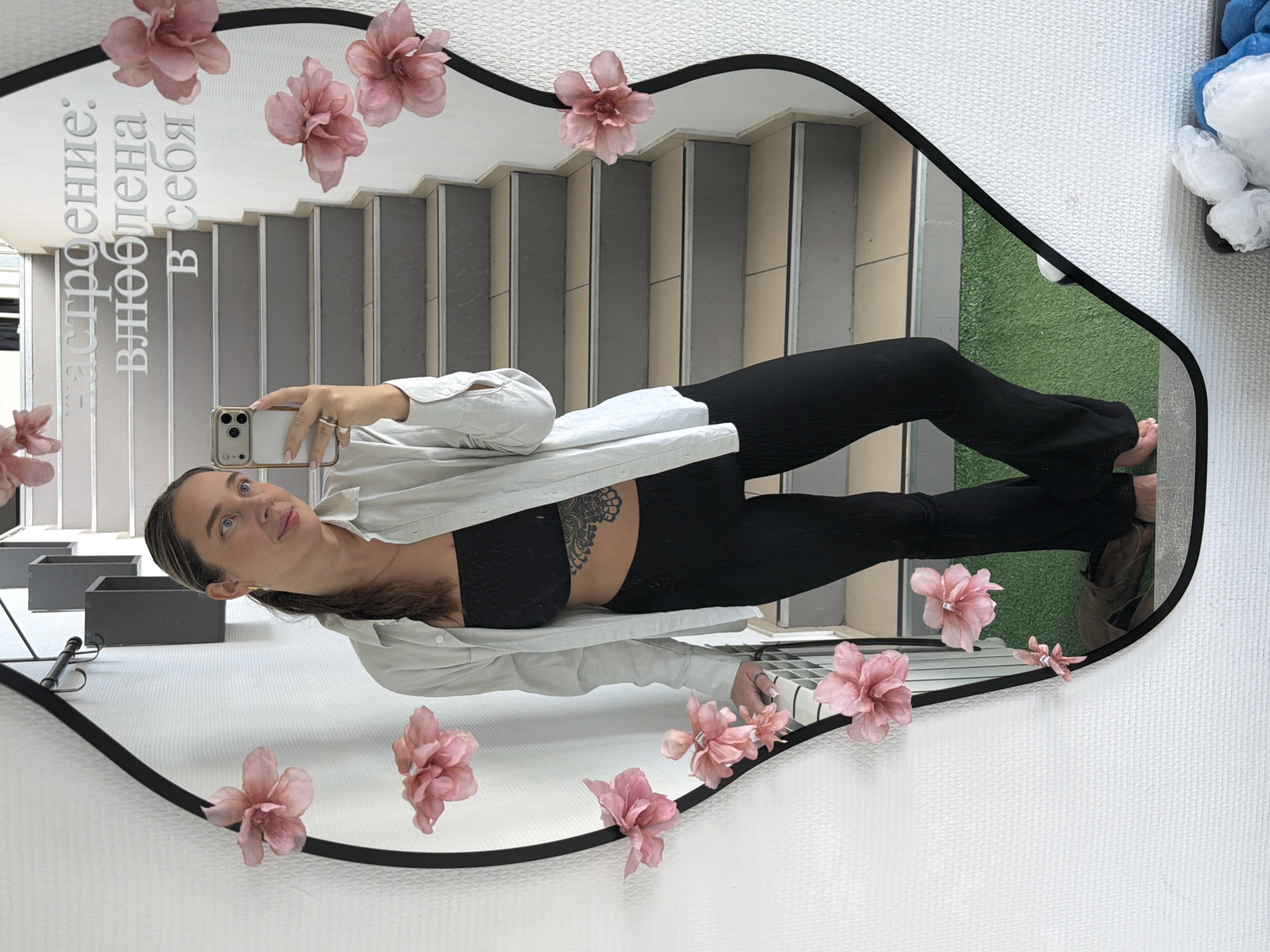 A woman in black leggings, a black top, and a light-colored, buttoned-down shirt is taking a selfie in a decorative mirror. The mirror, which is shaped like an irregular oval with a black border, is adorned with pink flowers. The reflection in the mirror includes a set of gray stairs and a patch of artificial green grass. There is text on the mirror in Cyrillic. The background suggests an interior location within a building, potentially in the city of Chaikovsky, Russia. A white, fluffy object can be seen in the upper right corner.