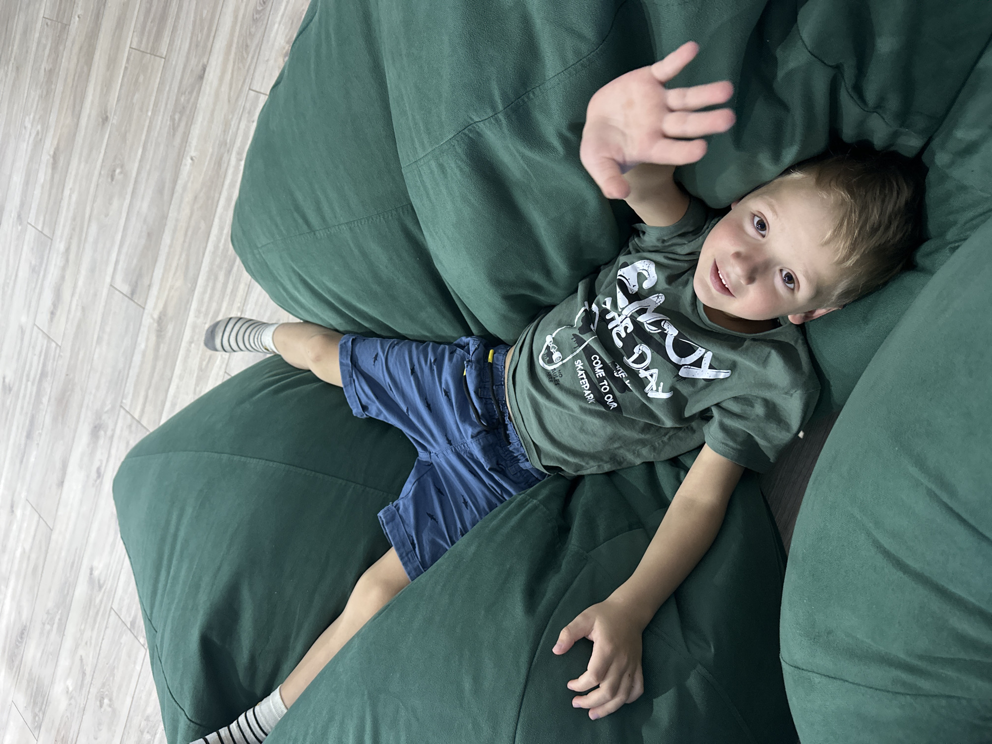 A young Caucasian boy lies upside down on a large, dark green beanbag chair. He is wearing a dark green t-shirt with white text and graphics, blue shorts with a shark pattern, and striped socks. His head is at the bottom of the frame, and his feet are at the top. The beanbag chair is on a light-colored wooden floor, likely in a residential setting. The boy appears to be looking up at the camera and has his right hand raised. The overall impression is of a relaxed and playful moment. The image was taken in Chaikovsky, Russia.