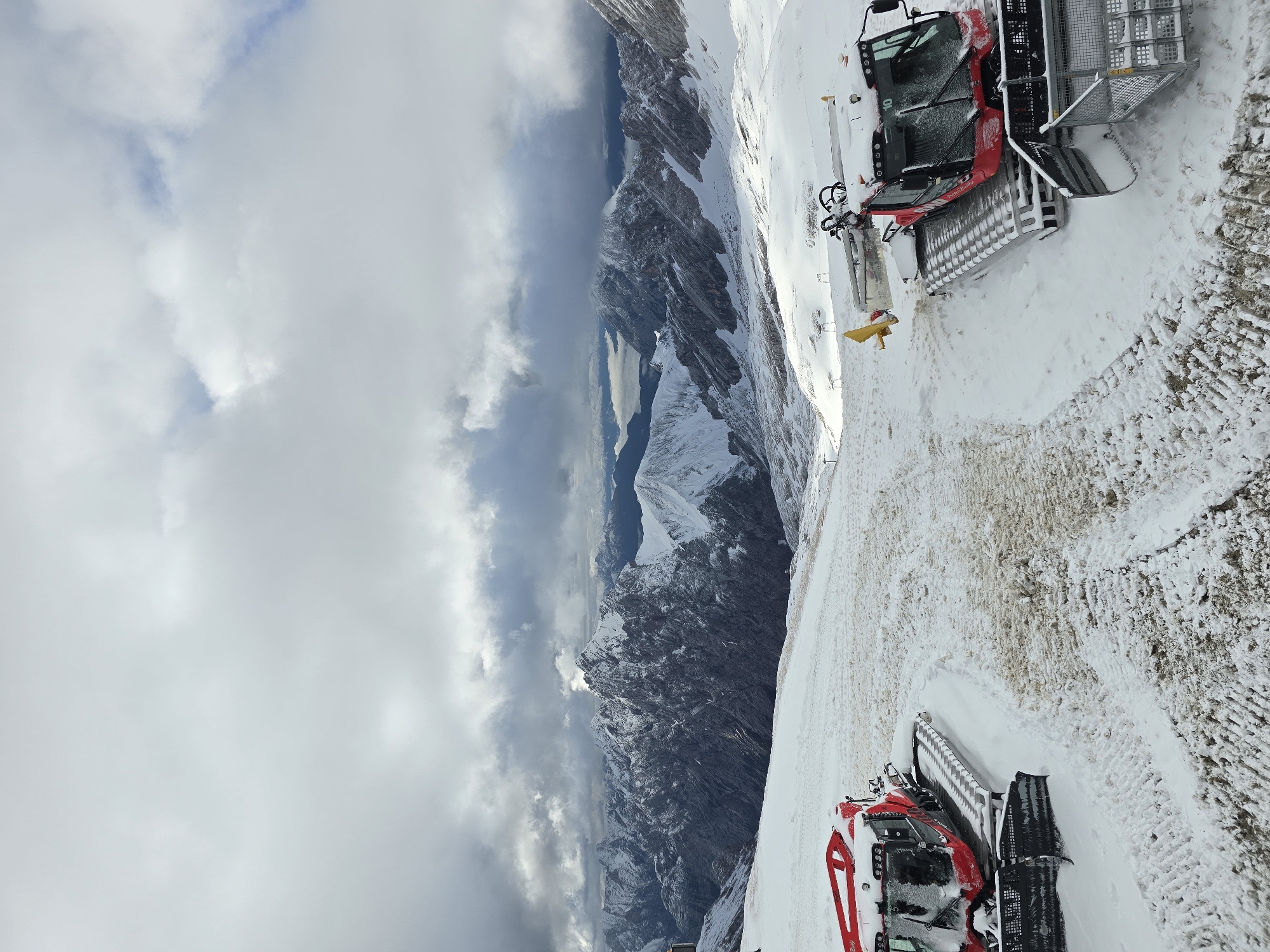 This image captures a panoramic view of a snowy mountain landscape, likely in Garmisch-Partenkirchen, Germany, given the presence of ski resort infrastructure. Two red snow groomer vehicles are parked on a snow-covered slope in the foreground. The vehicle on the left is positioned at an angle, with its front facing left and slightly away from the viewer. The vehicle on the right is positioned more towards the center right of the frame, with its front facing towards the viewer and slightly to the right. Both vehicles appear to be idle, covered in a light dusting of snow.
Behind the snow groomers, the terrain slopes downwards, showing tracks from previous grooming activities, indicating a ski slope or preparation area. In the mid-ground and background, a dramatic mountain range unfolds under a cloudy sky. The peaks are rugged and snow-capped, with stark contrasts between shadowed crevices and sunlit faces. Some of these mountains appear to be the Zugspitze massif, Germany's highest peak. The sky is filled with a mix of dark, heavy clouds and brighter, more diffused patches, suggesting a dynamic weather system. The overall impression is one of a remote, high-altitude winter environment.