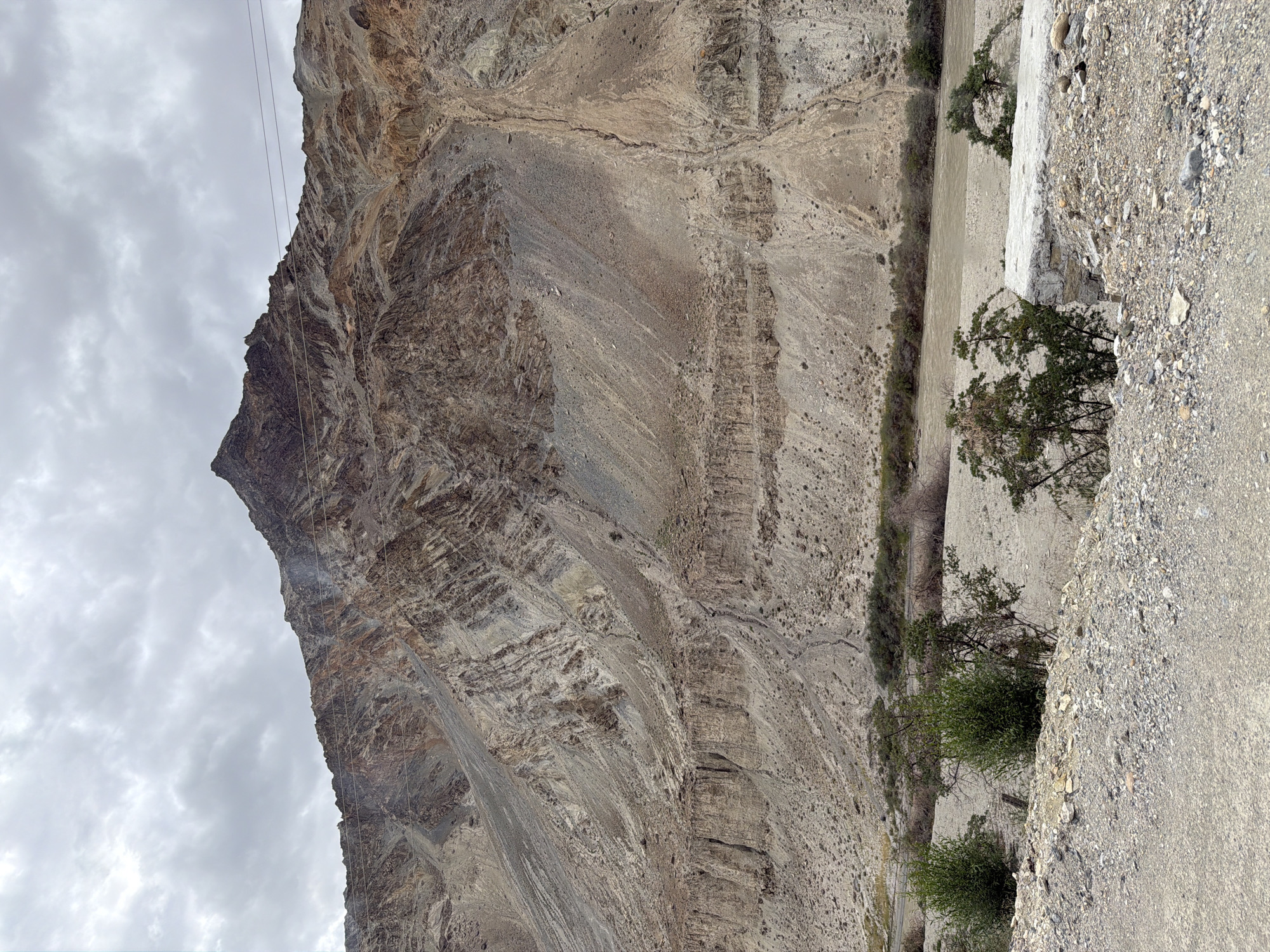 The image shows a mountainous landscape in the city of Chilling, India. The dominant feature is a large, barren mountain with distinct geological layers and striations, colored in shades of brown, beige, and gray. The mountain has a sharp peak. In the midground, a brown river flows through a wide, dry riverbed lined with sparse vegetation and small trees. The foreground consists of a gravelly, dusty terrain with scattered rocks and some more vegetation. The sky is overcast with gray clouds. There are no people or animals visible in the scene. Several utility wires are strung across the upper part of the mountain.