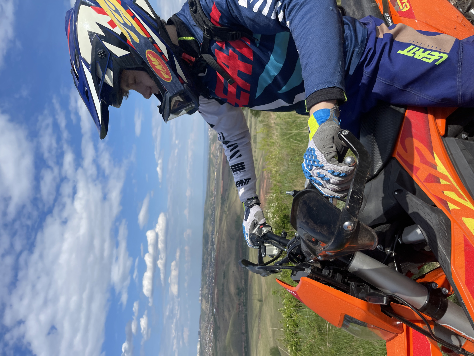 A person wearing a blue and red motorcycle suit, helmet, and gloves sits on an orange motorcycle. The person is holding the handlebars, looking downwards. The background shows a landscape with green fields, a town, and a blue sky with white clouds. The town is Солонцы, Russia.